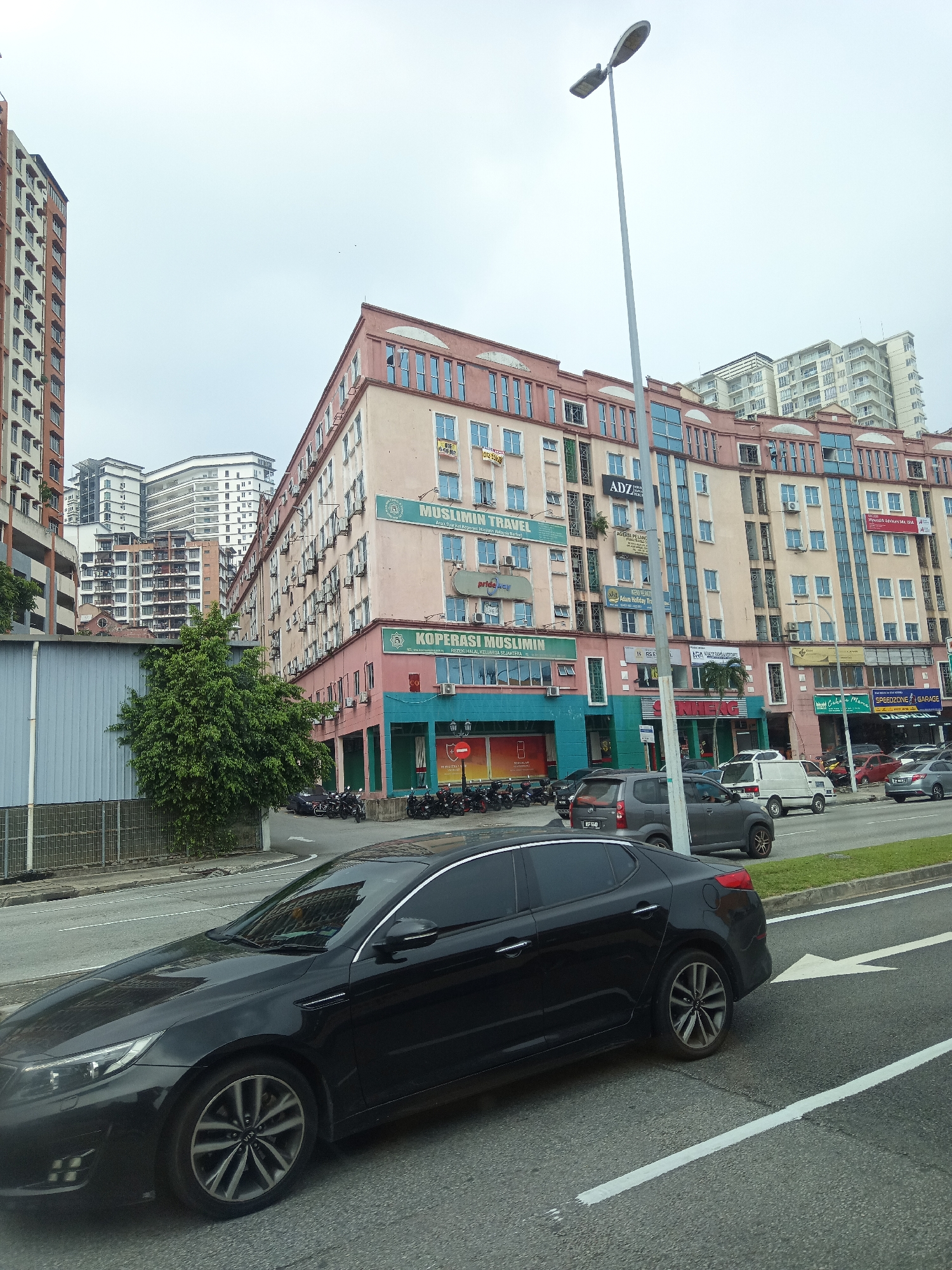 A black Kia Optima sedan is parked on the right side of a multi-lane road in Kuala Lumpur, Malaysia. The car is positioned in the foreground, partially obscuring the view of the street and surrounding buildings. The road is asphalt and appears to be in good condition, with visible white lane markings. In the background, a row of multi-story buildings lines the street, displaying various storefronts and office signs, including "MUSLIMIN TRAVEL" and "KOPERASI MUSLIMIN". The architecture of the buildings is diverse, with some featuring modern glass facades and others older, more traditional designs. A tall street lamp stands prominently on the right side of the image, casting a faint shadow on the road. The sky is overcast, suggesting a cloudy or hazy day. Several other vehicles, including a grey SUV and a white van, are visible further down the road. The general environment appears to be a commercial or urban area.