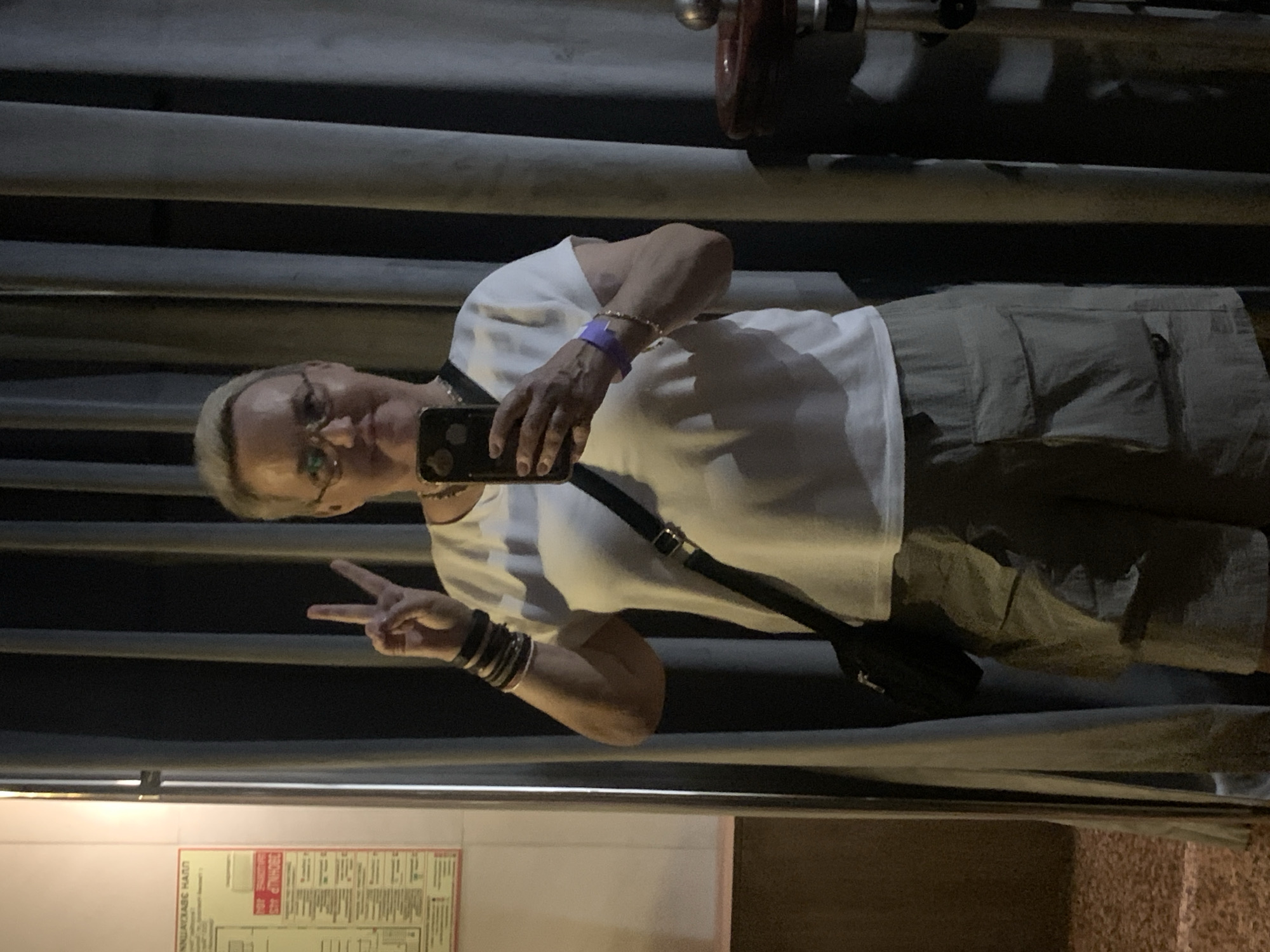The image shows a person with short blonde hair and glasses, wearing a white t-shirt and khaki shorts. They are holding a smartphone in their right hand, taking a selfie. The person is making a peace sign with their left hand. They are wearing several bracelets on their left wrist and a purple wristband on their right wrist. A black strap of a bag is visible across their chest. The background consists of horizontal, dark grey or black surfaces, possibly pipes or railings, with some lighter grey areas. In the lower part of the image, there is a yellow sign with text in red and black.
The time of day appears to be evening or night, given the low light conditions. The setting seems to be outdoors or in a space with industrial-like features. The person's expression is difficult to discern clearly due to the angle and lighting, but the peace sign suggests a casual or playful mood. The visible text on the yellow sign is in Russian. One prominent part of the text reads "ПОЖАРНАЯ БЕЗОПАСНОСТЬ" which translates to "FIRE SAFETY". Another section of text includes "ИНСТРУКЦИЯ" meaning "INSTRUCTION". The overall context suggests a public area, possibly a facility or an establishment in Nizhny Novgorod, Russia.
