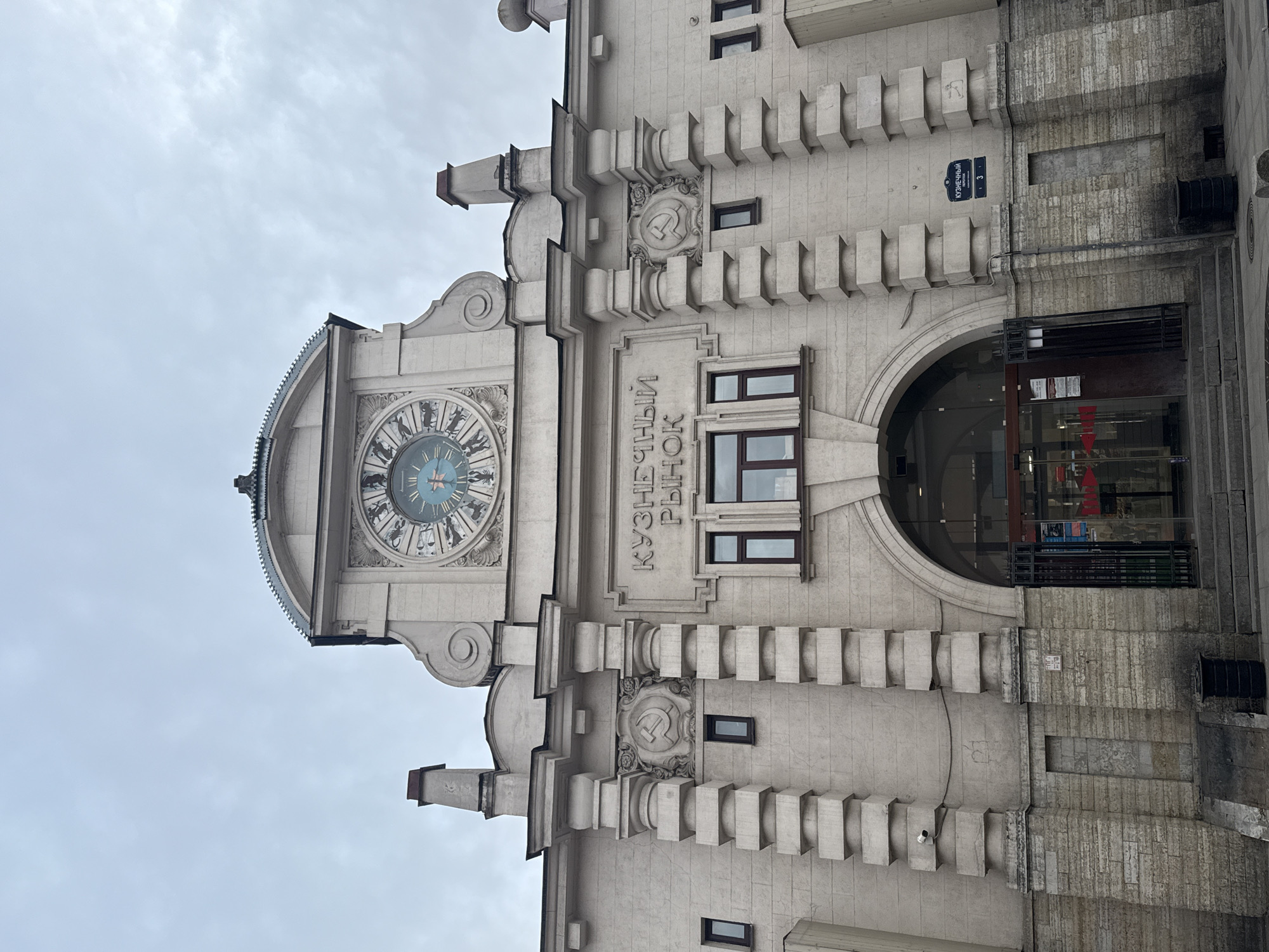 The image shows a close-up of the architectural facade of the Kuznetsov Market, a notable landmark in Saint Petersburg, Russia. The exterior is characterized by its elaborate Art Deco style, featuring a large clock at the top and Soviet-era symbols like the hammer and sickle. The facade also includes the market's name in Cyrillic ("Кузнечный рынок"). An entrance is visible on the right, with glass doors and reflections of the interior. The sky appears overcast. The building's exterior is made of light-colored stone with geometric details and vertical striations. A street sign with the name of the street and building number is affixed near the entrance.