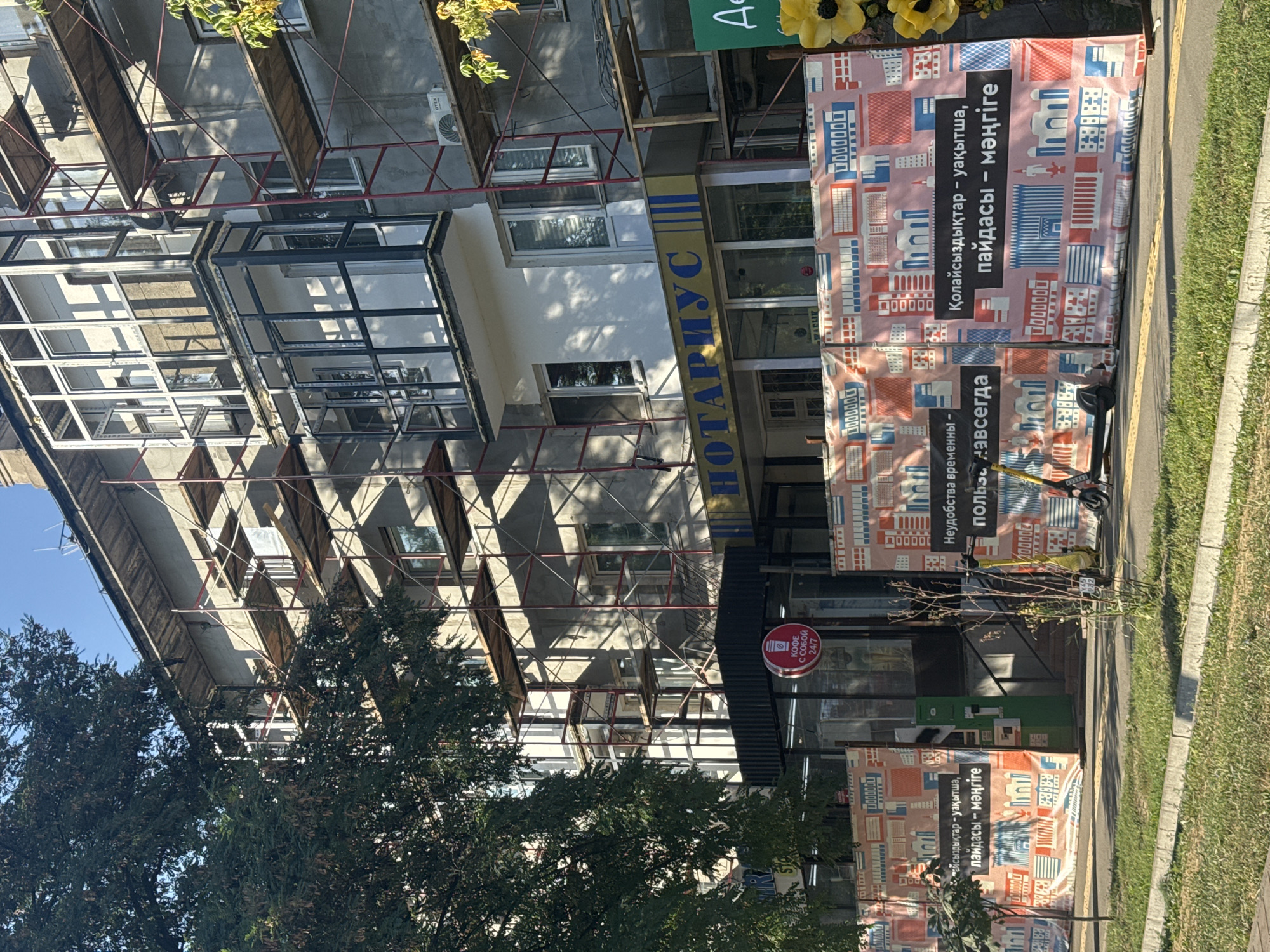 This is a medium shot of a building undergoing renovation in Almaty, Kazakhstan, viewed from across a street. The building is several stories tall, with scaffolding covering its facade. A large tree with dark green foliage is on the left side of the frame, partially obscuring the building. The building has several windows, some of which are covered or reflective. There is a prominent sign with yellow and blue lettering that reads "НОТАРИУС" (Notary) above a storefront. In front of the building, a banner with a repeating pattern of stylized buildings and geometric shapes is hung, with some text in Kazakh. Two electric scooters are parked in front of the banner. The environment is urban, with a sidewalk and a grassy median in the foreground. The sky is clear and blue.