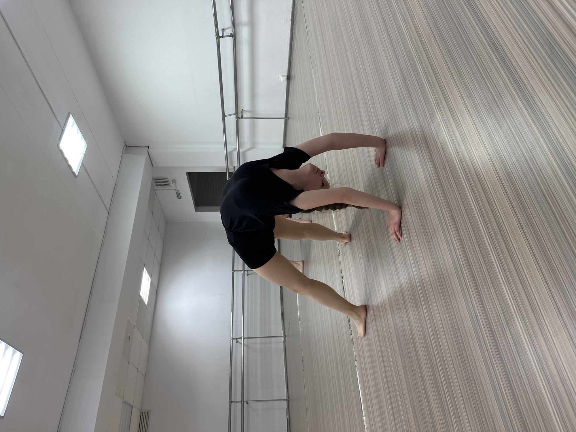 A woman in a black shirt and black shorts is performing a backbend on a floor in a dance studio. Her hands and feet are flat on the floor for support. The studio has gray-toned floor panels, white walls, and a ballet barre fixed to the wall. There are fluorescent lights on the ceiling. The location is Votkinsk, Russia.