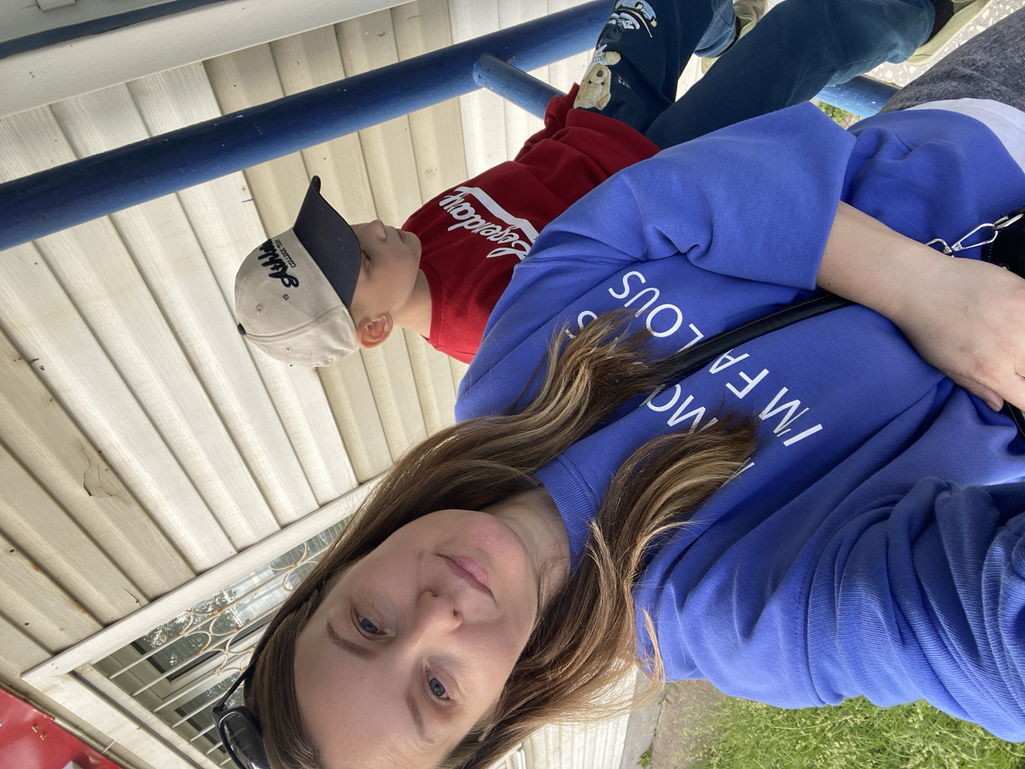 This image, taken in Chaikovsky, Russia, features a woman and a child outdoors. The perspective is a selfie, with the woman holding the camera and appearing in the foreground.
The woman has long, wavy brown hair and blue eyes, looking directly at the camera with a neutral to slight smiling expression. She is wearing a vibrant blue crewneck sweatshirt with white text on the chest that reads "I'M FABULOUS". Sunglasses are tucked into the neckline of her shirt, and a black strap, possibly from a bag or glasses, is visible over her right shoulder.
Behind and slightly to her left, a child is visible, facing away from the camera. The child wears a red long-sleeved shirt with the word "Legendary" in white lettering across the chest. They also have on dark pants adorned with cartoon teddy bear graphics and a light-colored baseball cap with a dark brim. Text on the cap partially visible includes "Athletic College 19xx" (the last two digits are obscured). The child's gaze is directed to the left, away from the camera.
The setting appears to be an exterior location next to a building. A horizontal blue metal railing spans across the middle background, separating the subjects. Behind the railing, the building features light-colored, horizontal siding. A small window with decorative metal bars is visible to the left of the woman's head. Green grass is present in the lower right portion of the frame, and light pavement or ground is visible elsewhere. The lighting suggests it is daytime, possibly under an overcast sky, creating soft, even illumination.