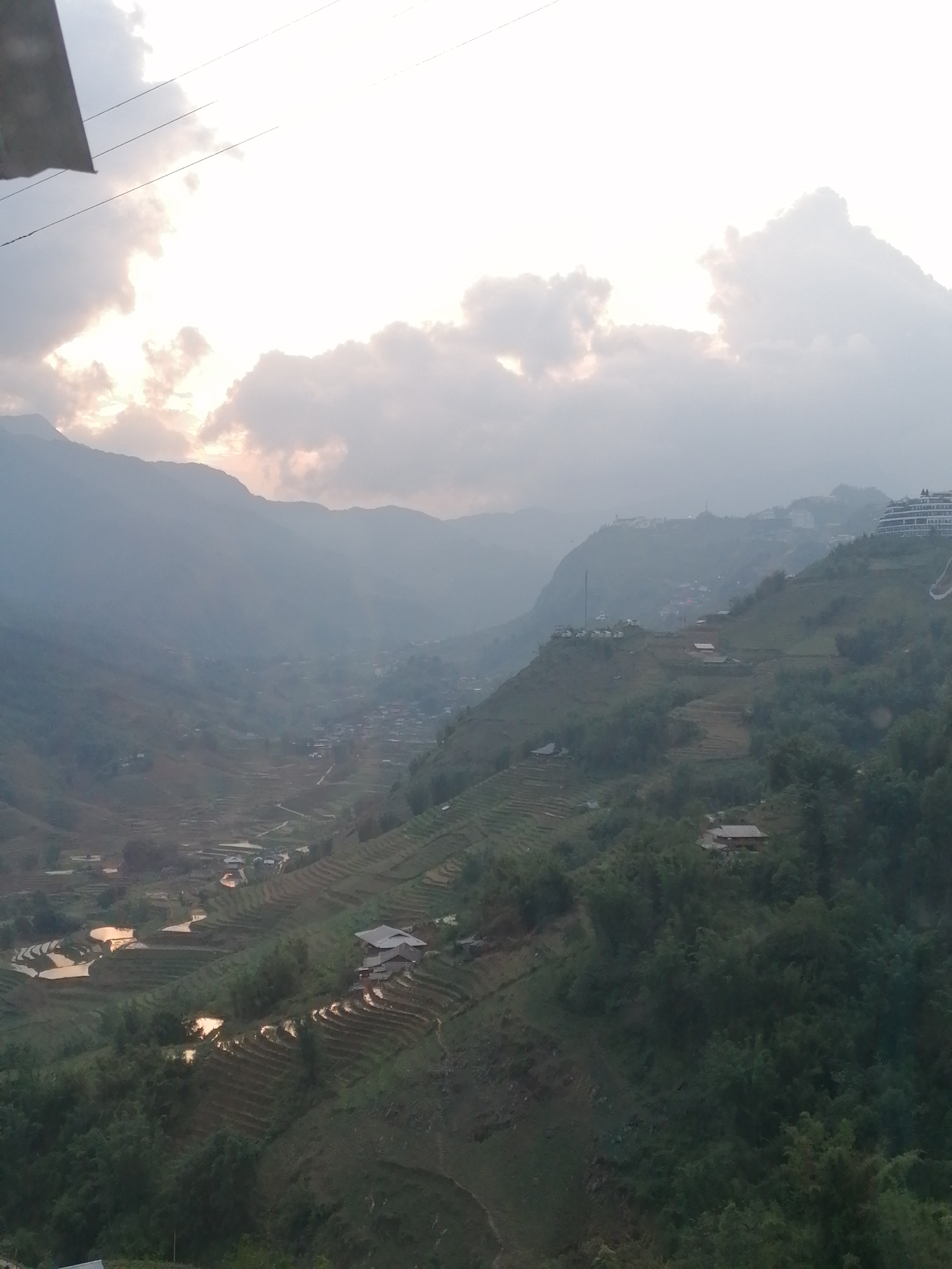 This panoramic image presents a vast, hazy landscape of a mountainous valley in Sa Pa Ward, Vietnam, captured during either sunrise or sunset. The sky is predominantly cloudy, with soft, diffused light filtering through, casting warm, pale golden and pink tones on the clouds. Power lines are faintly visible against the bright sky in the upper left corner.
The scene is dominated by a deep valley flanked by multiple layers of mountains, which appear progressively lighter and bluer due to atmospheric haze. The hillsides are intricately carved into extensive terraced fields, characteristic of rice cultivation in the region. Many of these terraces, particularly in the mid-ground, are filled with water, reflecting the bright sky and creating luminous patches within the verdant and earthy landscape.
Scattered across the valley floor and along the lower mountain slopes are numerous small houses and clusters of buildings, forming villages. On a distant ridge to the right, more modern development is visible, including a multi-story building complex with a distinctive curved design and a tall communication antenna or mast. Lush green trees and vegetation cover the non-cultivated areas and steeper inclines.
No explicit human activity is depicted, but the presence of the terraced fields and scattered dwellings indicates an agricultural and settled community. The time of day is suggested by the soft, indirect lighting and warm hues in the clouds. No visible text is present in the image. The overall impression is one of tranquil natural beauty and traditional rural life within a dramatic mountainous setting.