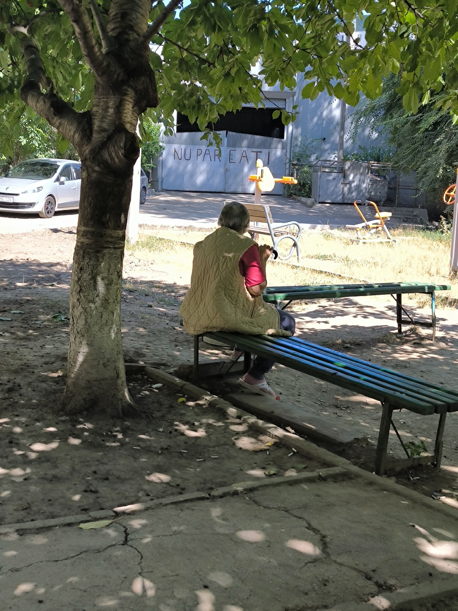 An elderly person with gray hair, wearing a light brown vest over a red shirt and dark pants, sits on a green slatted bench. Their back is to the camera, and they are holding a black object in their right hand, possibly a phone or a small tool. They are wearing white open-toed sandals with orange accents. The bench is placed on a paved area, with another green slatted bench parallel to it in the foreground, which appears to be painted with blue and green stripes. Behind the benches, there is a tree with green foliage casting shadows on the ground. In the background, a silver car is parked on the left, and a large, gray metal gate with the text "NU PAR EATI" painted in a darker color is visible. There are also children's playground equipment, including a yellow and orange swing set, and a white and orange spinning toy, further in the background. The environment suggests an outdoor public space, likely a park or a courtyard in a residential area within Chișinău, Moldova. The ground is a mix of dirt, gravel, and cracked concrete. Several cigarette butts are scattered on the ground. The overall scene depicts a moment of rest in a somewhat neglected outdoor setting.