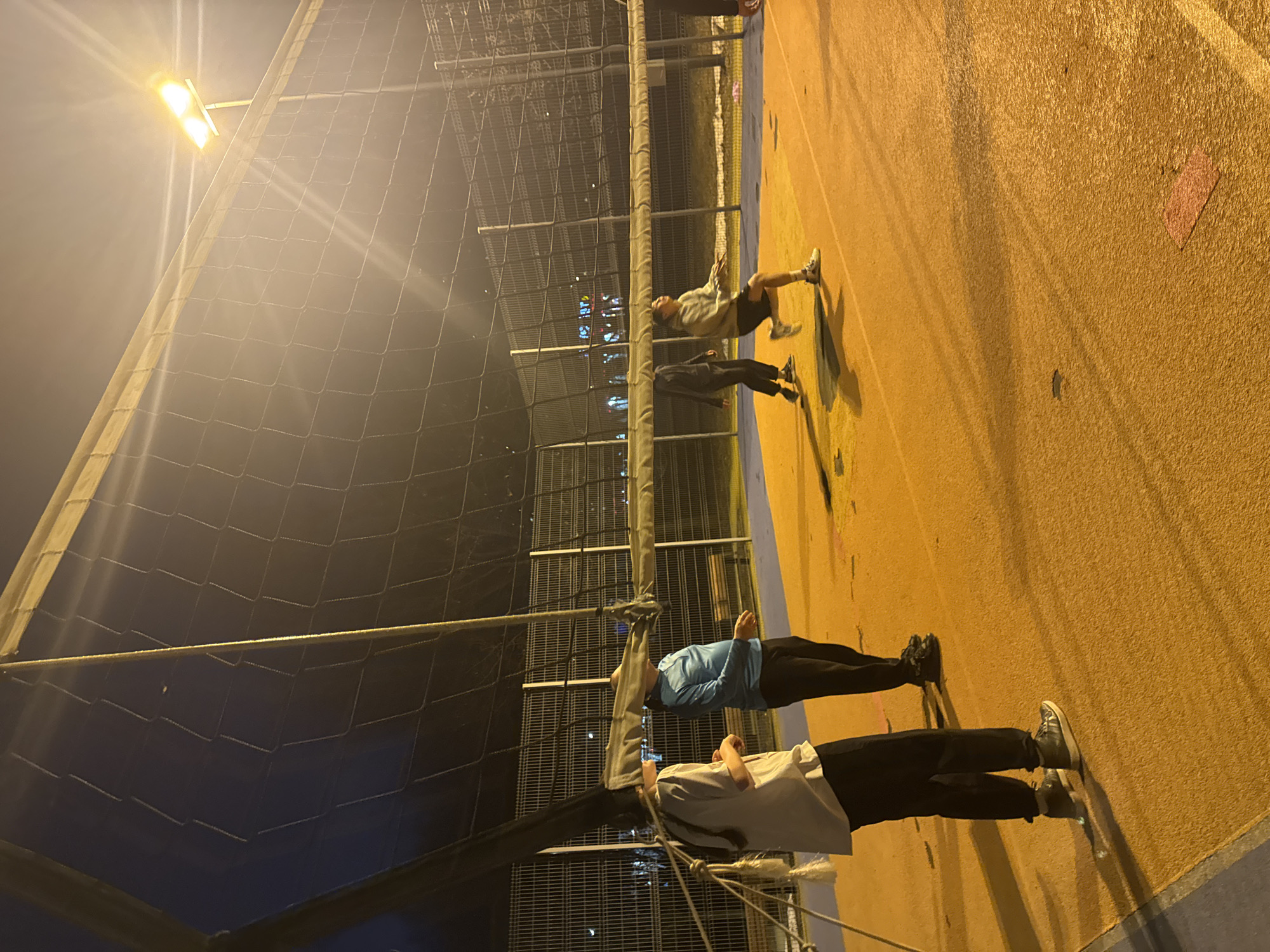 The image shows a nighttime scene on what appears to be a sports court or recreational area. There are at least four people present, positioned on the right side of the image, parallel to a netting structure. The netting separates the playing area from what looks like a fence. The background displays out-of-focus lights, indicative of a cityscape. The area is illuminated by artificial lights, casting shadows on the ground. Based on the overall style and available features, it may be a Moscow sports court.