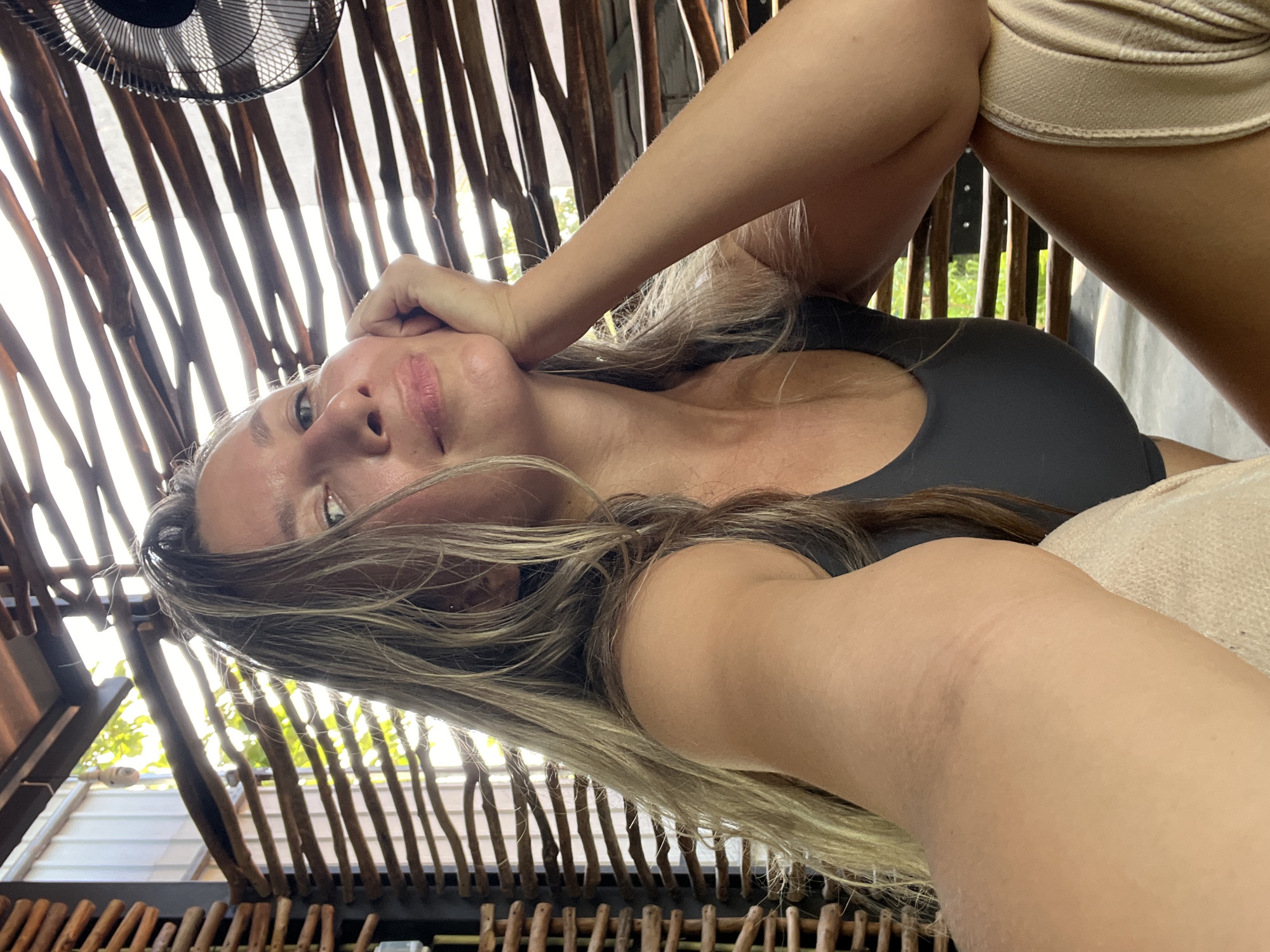 The image is a selfie taken by a woman with long blonde hair and light skin. She is wearing a dark gray tank top and is leaning on her hand, with her elbow propped on her knee. She is looking at the camera with a slight smile. The background consists of a wooden slatted structure, with sunlight filtering through it, creating a striped effect. A portion of a fan is visible in the upper right corner of the image. The woman appears to be in an outdoor, shaded area. The location is identified as Weligama, Sri Lanka.