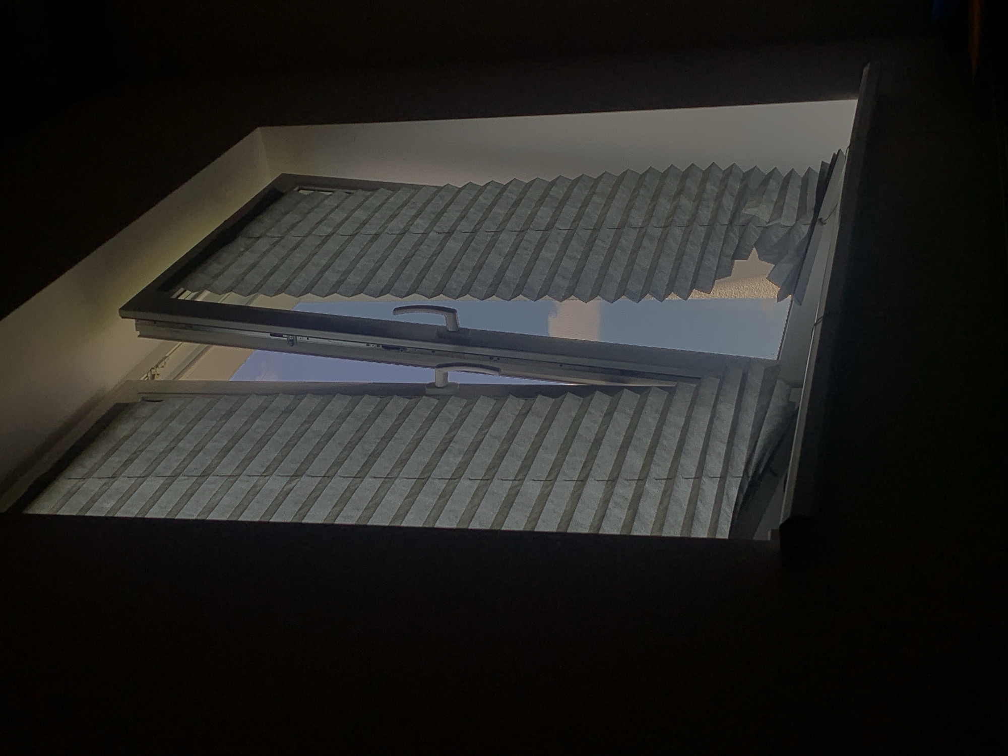 The image shows a partially opened window with pleated blinds. The window frame is dark, and the blinds are a light grey color, with some sections appearing creased or damaged. Through the open part of the window, a bright blue sky with some white clouds is visible. The scene is framed by dark, shadowy areas on either side, suggesting the view is from inside a dimly lit room looking out. The location is specified as Minsk, Belarus.