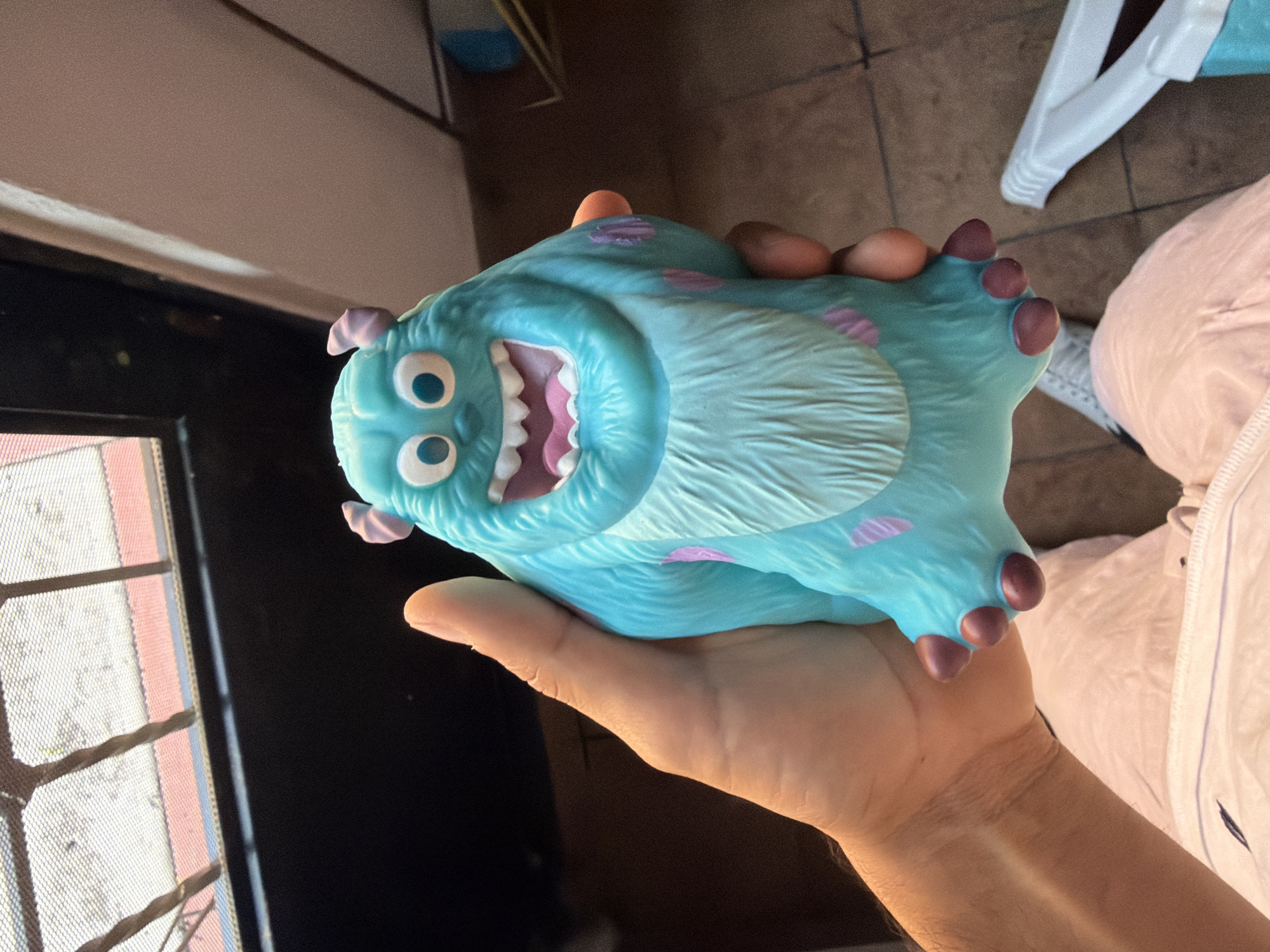 A person is holding a blue Sully toy from "Monsters, Inc." in their right hand. The toy has a large mouth open, purple spots, and horns. The background includes a door with a screened window to the left, a tiled floor, a white chair, and a person wearing white clothing to the right. Based on the metadata, the location is Reforma, Mexico.
