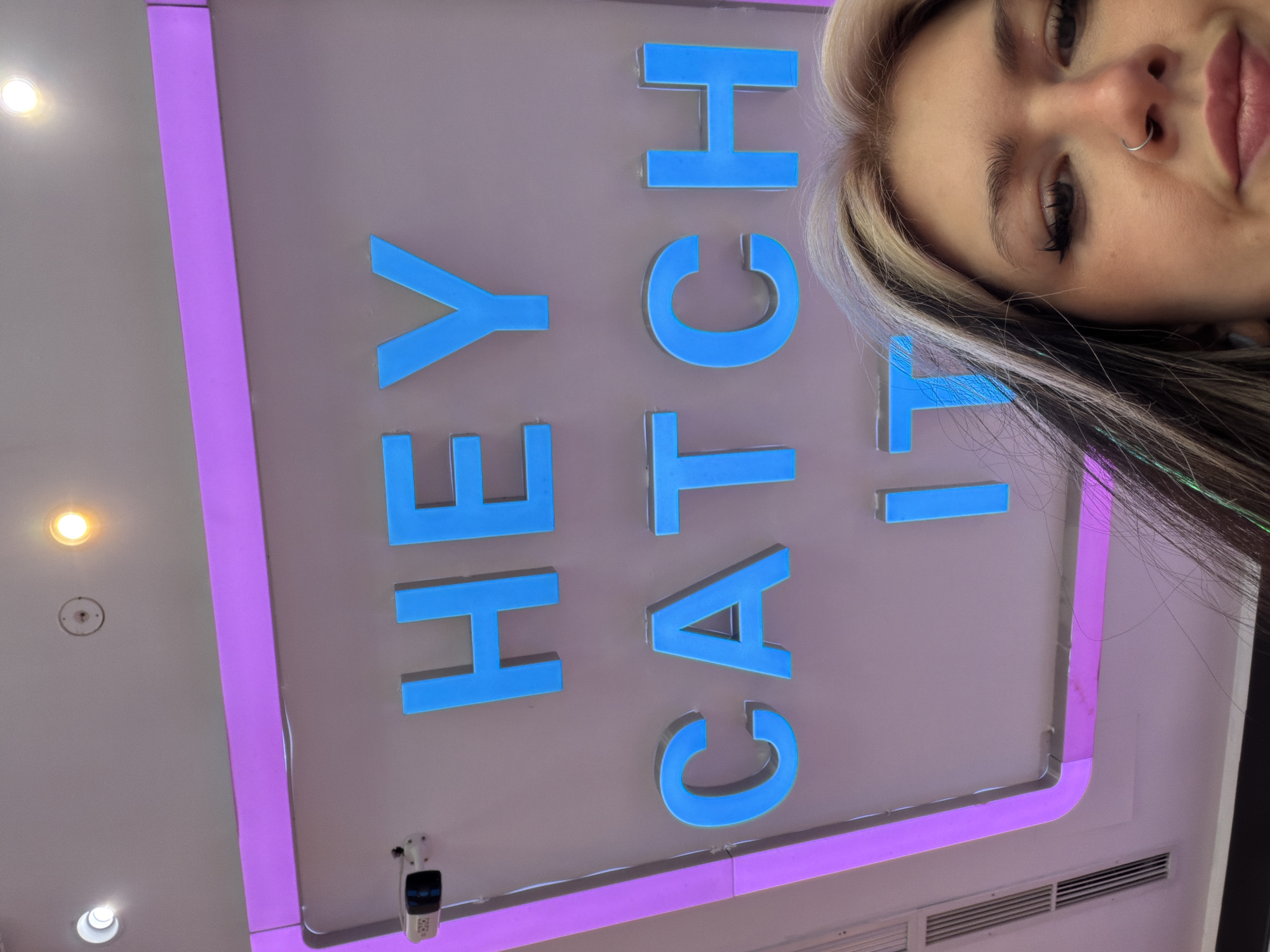 A low-angle shot shows a young woman with bleached blonde and dark hair, a nose ring, and makeup, partially obscuring a sign with the words "HEY CATCH IT!" written in large, blue capital letters. The sign is framed by a purple border. The location appears to be indoors, possibly a retail space, as indicated by the ceiling with recessed lighting and ventilation grilles. A security camera is mounted near the bottom of the sign. The exact location in Palilula Urban Municipality, Serbia cannot be determined from the image alone.