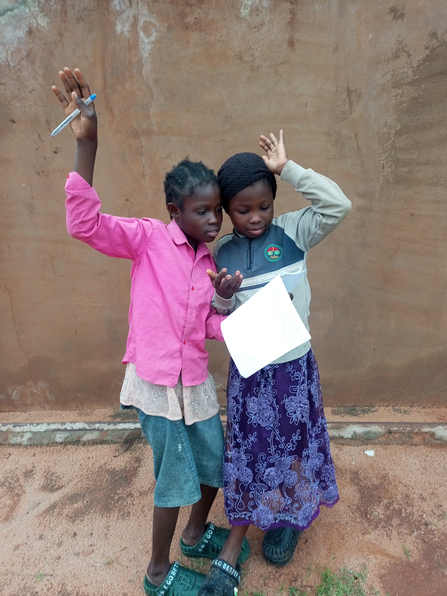 Two young girls, appearing to be of African descent, are standing outdoors in front of a textured, earth-toned wall. The girl on the left, wearing a bright pink collared shirt over a light-colored, possibly lace-trimmed skirt and denim shorts, holds a blue pen aloft in her right hand, her palm facing upwards. Her left hand is extended towards the girl on the right. The girl on the right is wearing a dark beanie, a light-colored long-sleeved shirt with a dark grey and blue panel on the front featuring a circular emblem, and a dark purple skirt with a floral lace overlay. She is holding a white piece of paper in her hands, looking down at it. Both girls are wearing green slide sandals. The ground beneath them is composed of reddish-brown dirt with sparse patches of green grass. The overall setting suggests an outdoor environment in a warm climate. The specific location in the city of Tempure, Nigeria, is not discernible from the image alone.