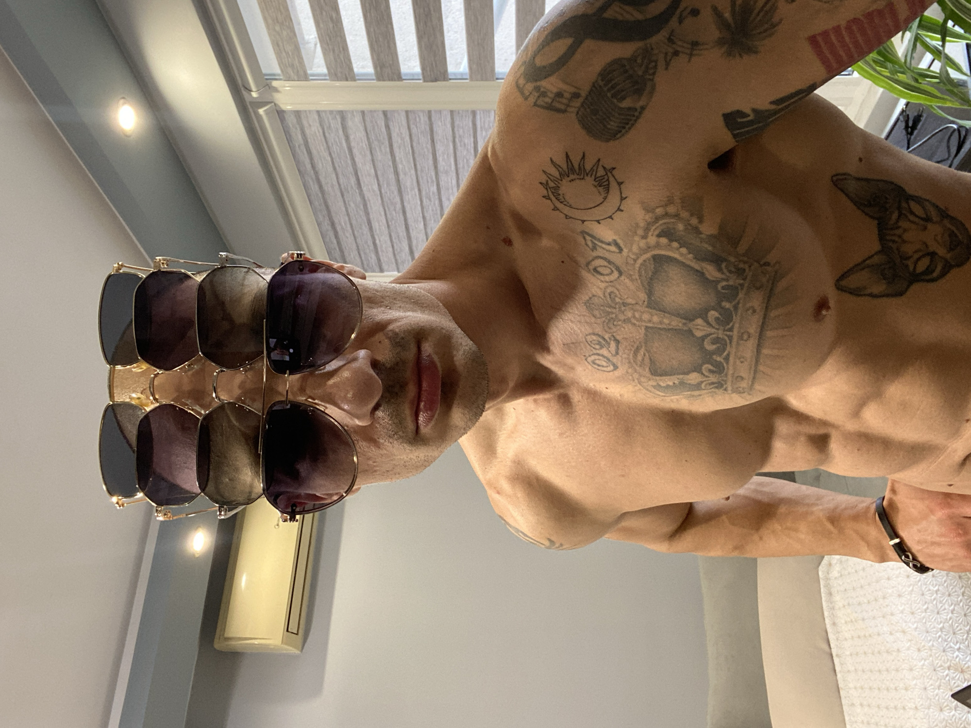 A Caucasian man stands indoors. The man's upper torso is visible, and he is not wearing a shirt. He has multiple tattoos on his shoulders and chest including a crown, microphone, sun-like symbol, and other illegible designs. He is wearing multiple pairs of sunglasses on his head, stacked on top of one another. He has short dark hair. The background shows a closed window blind and a neutral gray wall. A vertical perfume bottle sits on a shelf in the background. There is no indication that this photo was taken in the city of Pleven, Bulgaria.