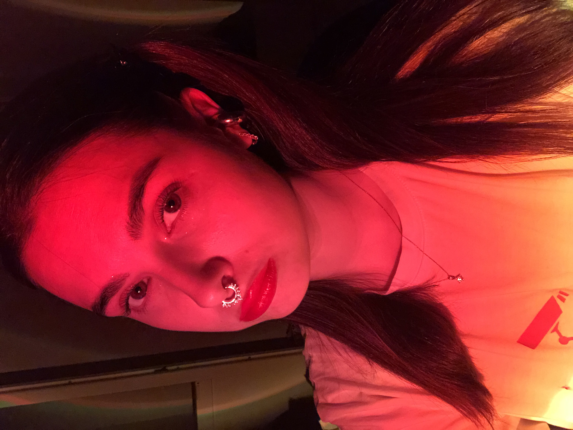 A person with long, dark hair, wearing a nose ring and earrings, and red lipstick is illuminated by red and orange light. The person is wearing a light-colored shirt, and a necklace is visible. A logo is faintly visible on the shirt. The scene appears indoors, possibly near a monitor. The city and country are irrelevant to the visual information available in the image.