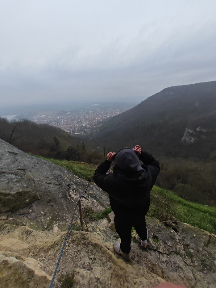 A single individual stands on a rugged, light-colored rocky outcrop, viewed from behind, facing away from the camera. The person is dressed in a dark hooded jacket, with the hood pulled over their head, and dark pants, with light-colored footwear. Both hands are raised, positioned near the hood, possibly adjusting it.
The scene provides a panoramic view overlooking the city of Shumen, Bulgaria, situated in a valley below. The immediate foreground features irregular rock formations and patches of green grass. A blue rope or cable is secured to the rock face on the left, running downwards, with visible metal anchors embedded in the stone, indicative of a via ferrata or secured path.
Beyond the individual and the immediate rocky terrain, steep, dark-forested slopes descend into the valley. The city of Shumen occupies the valley floor, presenting a dense urban landscape of buildings under a flat, uniformly overcast grey sky. Distant plains extend beyond the city, partially obscured by atmospheric haze. The environment is characterized by natural mountainous terrain overlooking an urban settlement.