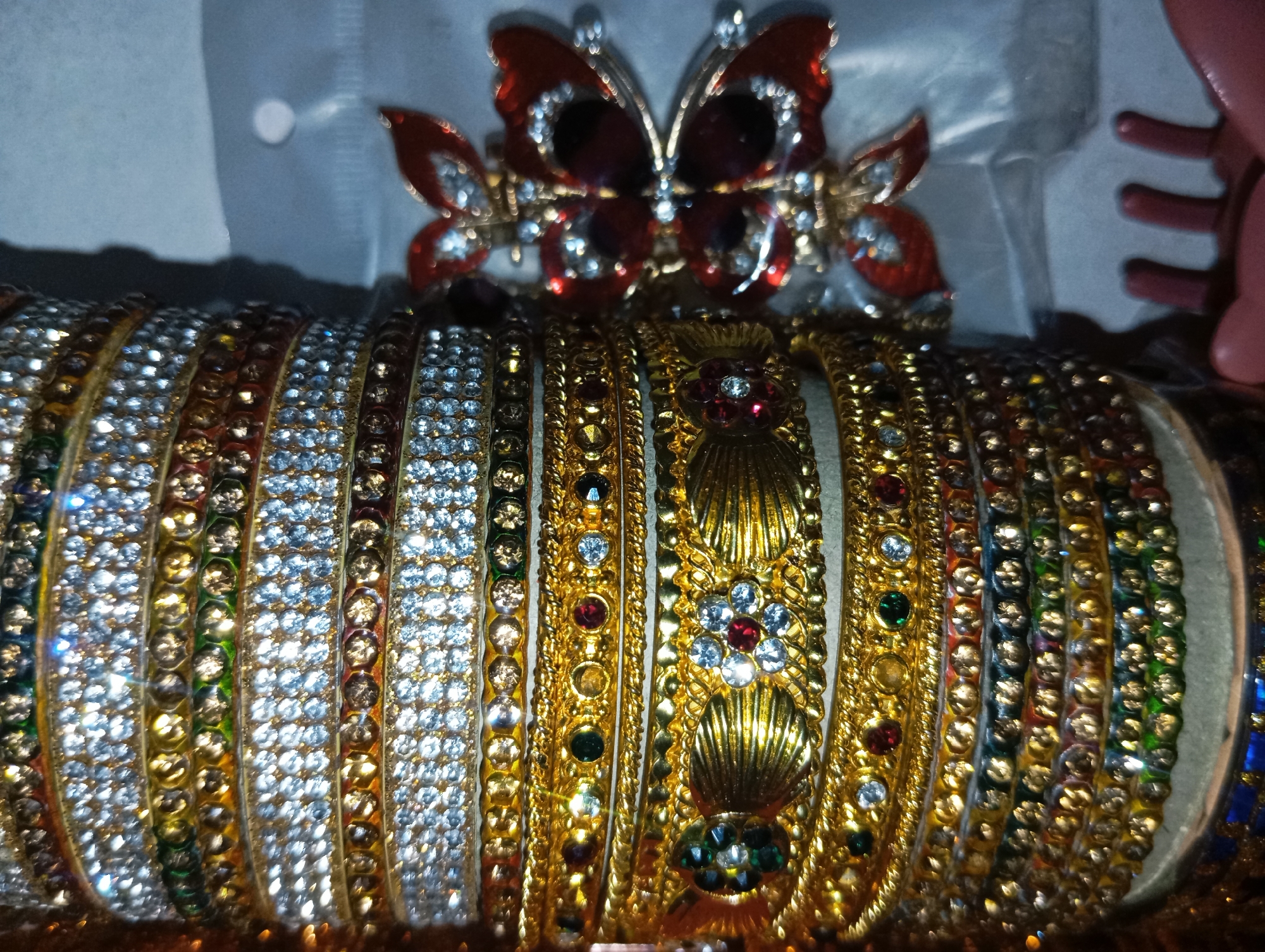 This is a close-up, eye-level, medium shot of several gold and silver bangles adorned with colorful rhinestones. The bangles are arranged in a row, with the gold bangles featuring intricate embossed designs and some also containing red, green, and clear gemstones. The silver bangles are densely covered in clear rhinestones. A red and gold butterfly hair clip is visible at the top of the frame, positioned behind the bangles. The background is a soft, out-of-focus light gray. The lighting is somewhat dim, with bright highlights reflecting off the gemstones and metal surfaces. The image appears to be taken indoors, possibly in a display case or on a surface. No people, text, or specific location cues are visible. The overall impression is one of decorative jewelry.