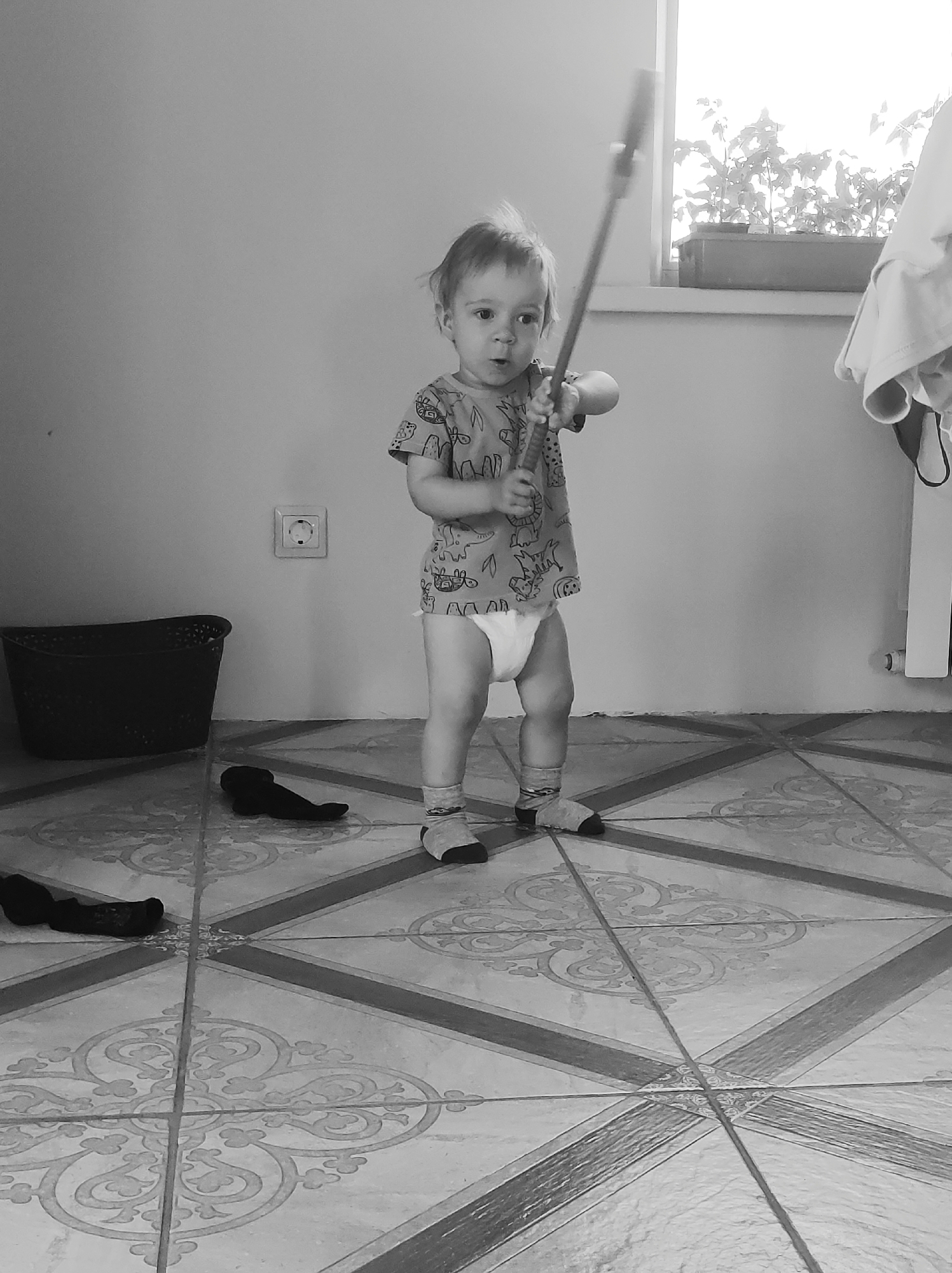 A black and white image depicts a baby standing on a patterned tile floor. The baby is wearing a printed shirt, diaper, and socks. The baby is holding a mop handle. A black basket and a pair of shoes sit on the floor. An electrical outlet is visible on the wall. A window with plants is in the background.
The location is not in the city of Фоки, Russia.