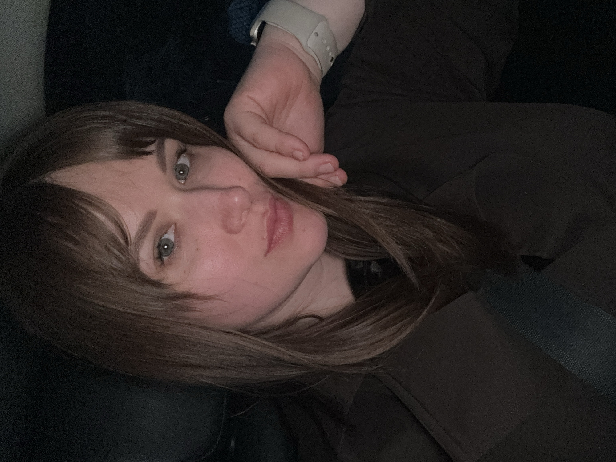 A woman with shoulder-length brown hair and bangs is reclining on a dark surface, possibly a seat, wearing a brown top. She has light skin, green eyes, and appears to be wearing a light-colored smartwatch on her left wrist. Her left hand is near her face. A dark seatbelt is visible across her body. The location in the city of Chaikovsky, Russia, cannot be determined from this image.