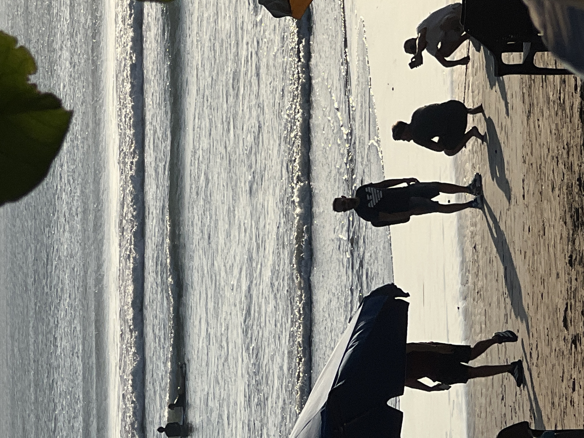 In Kuta, Indonesia, a group of people are gathered on a beach with the ocean and waves in the background. In the foreground, a dark blue umbrella is partially visible, casting a shadow on the sand. To the right of the umbrella, a person is walking away from the camera, wearing a dark t-shirt and shorts. Further down the beach, three people are interacting: one person stands looking towards the water, another is crouched down and appears to be taking a photo with a phone, and a third person is also crouched. In the middle ground, several people are in the water, some on surfboards. The sun is shining, creating bright reflections on the water and long shadows on the sand.