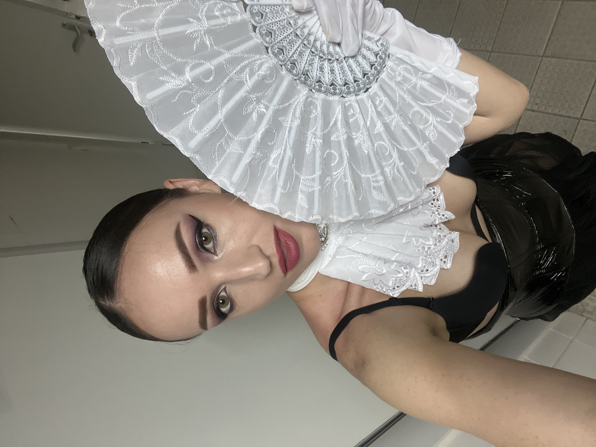A woman with dark hair pulled back from her face and wearing dramatic purple eye makeup and red lipstick poses for a selfie. She is holding a large white fan adorned with intricate embroidery and decorative metalwork in front of her face, obscuring part of it. She is also wearing a white, ruffled, lace collar, a black bra, and a shiny black skirt. The background appears to be an indoor setting with neutral-colored walls and tiled flooring.