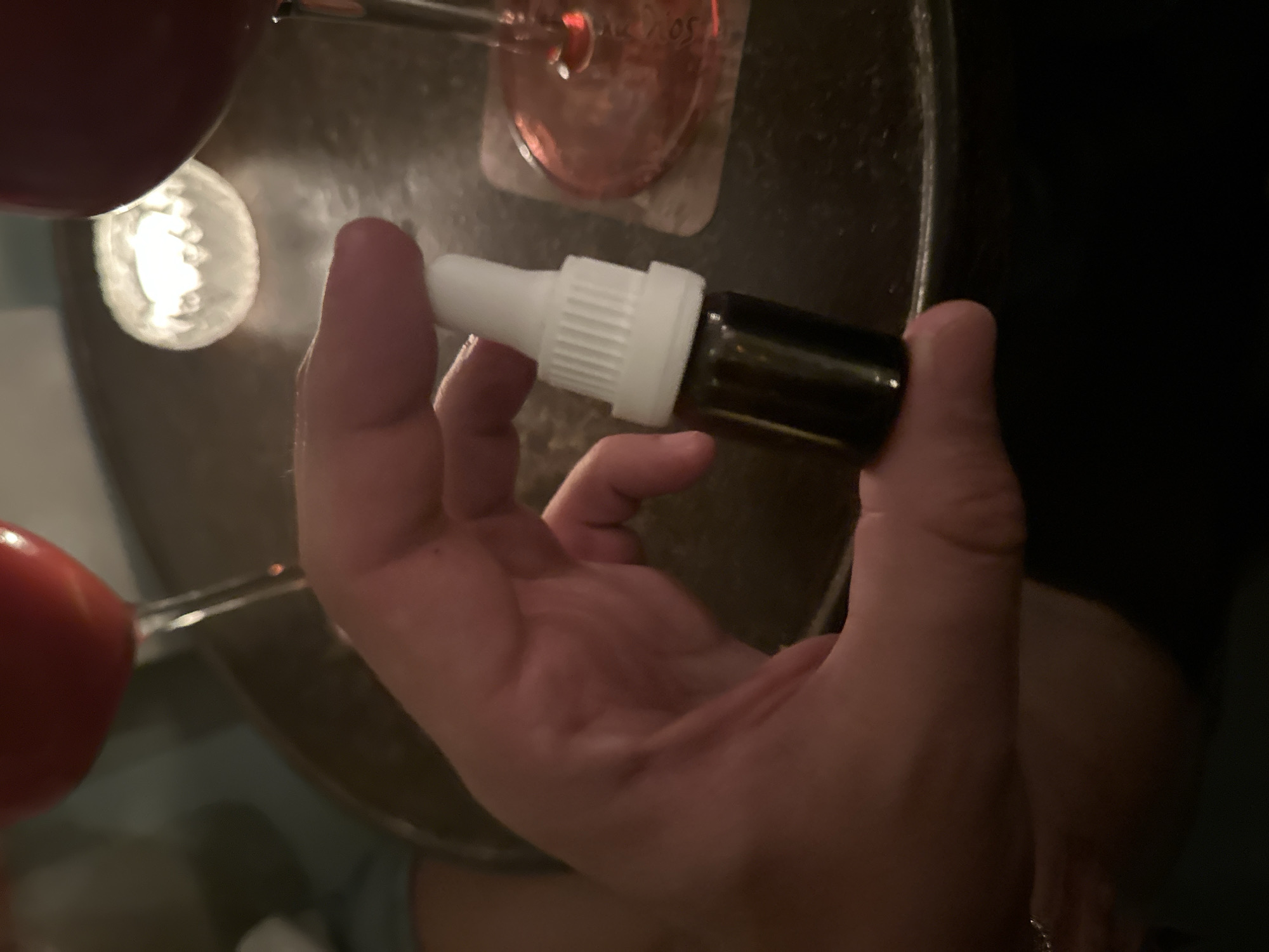 A hand holds a small, brown glass bottle with a white dropper cap. The bottle is positioned against a dark background. Out of focus elements include an out-of-focus object on the left side of the frame and what appears to be parts of glasses and a coaster on a surface. The overall lighting is dim. The location is unknown.