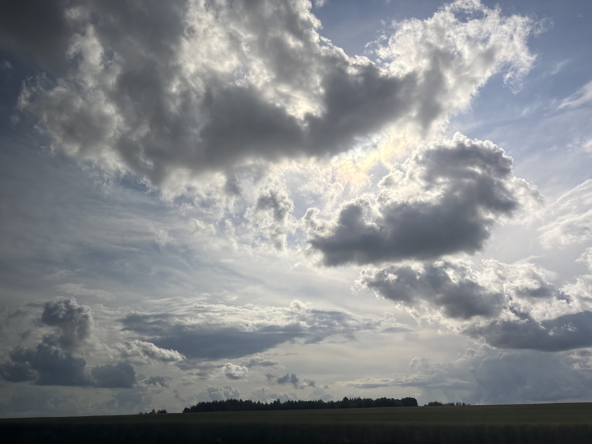 The image captures a dramatic sky over a rural landscape in the Senicki rural council, Belarus. Dark, voluminous storm clouds dominate the upper portion of the frame, interspersed with patches of brighter sky and beams of sunlight that create a sense of diffused light and possibly a rainbow effect. Below the sky, a dark, silhouetted treeline stretches horizontally across the middle ground, separating the sky from a dark foreground. The foreground appears to be a field, rendered in deep shadow. The overall impression is one of an approaching storm or the aftermath of one, with a strong contrast between the turbulent sky and the somber earth. There are no people or discernible structures visible in the image.