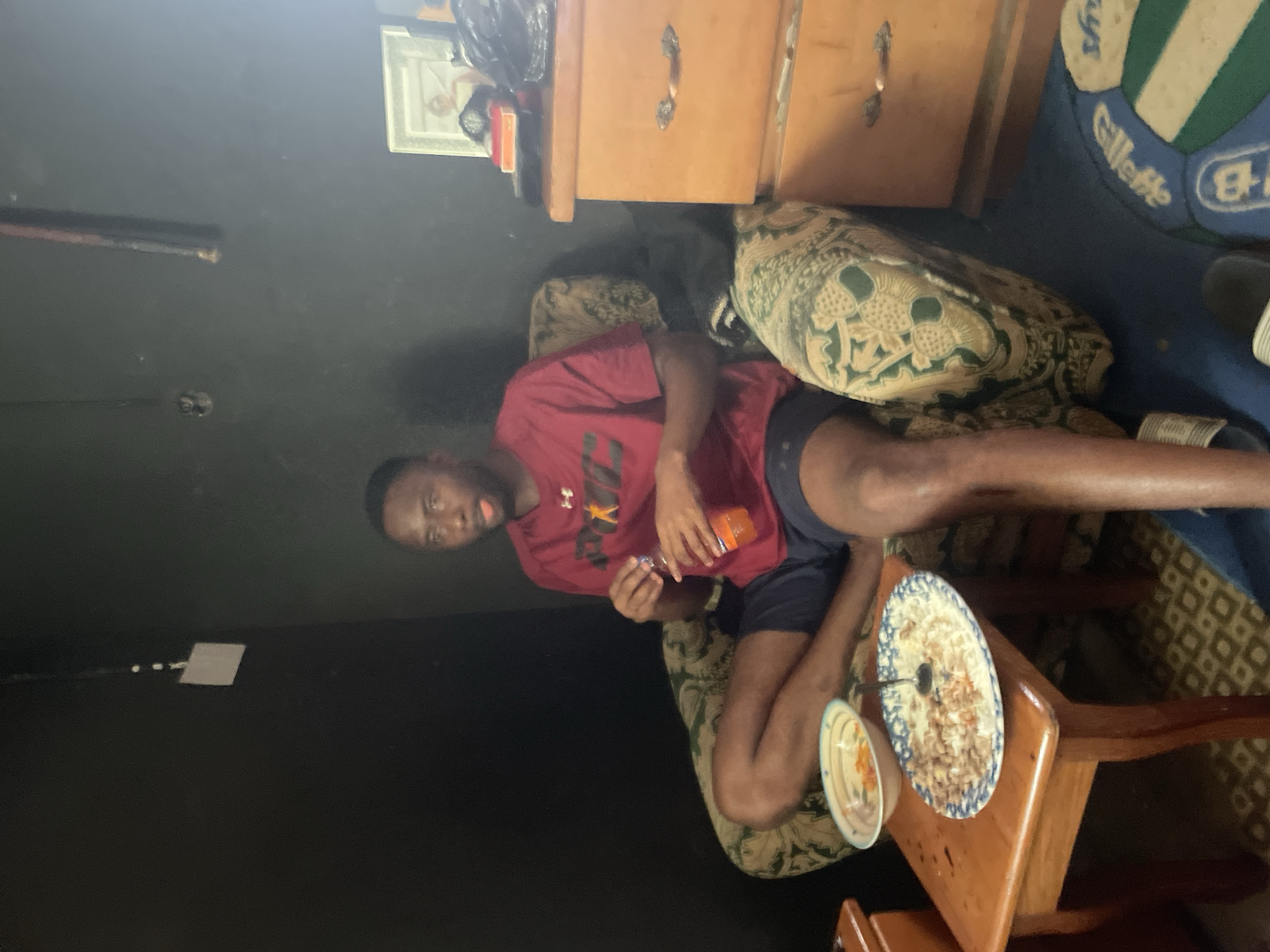A Black male sits in a patterned armchair, holding an orange beverage bottle and looking to his right. He is wearing a maroon t-shirt with a yellow star logo and dark blue shorts. In front of him, on a wooden side table, are two plates of food, one containing rice and meat, the other a lighter-colored food with some orange pieces. To his right, a light-colored wooden dresser with three drawers stands against a dark wall. A framed picture sits on top of the dresser. The room appears to be indoors.