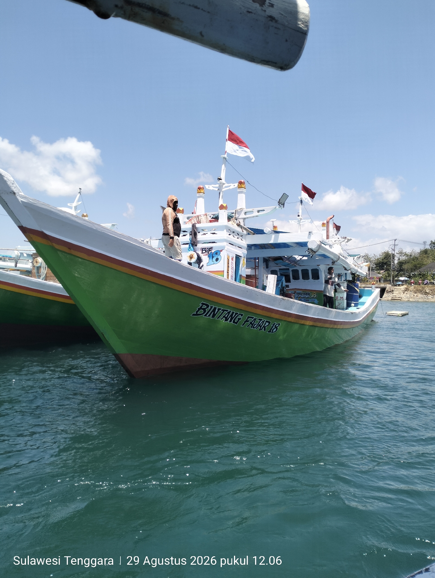 A fishing boat with the name "BINTANG FAJAR 18" is docked in the water, with a second boat partially visible behind it. The primary boat has a green hull and a white superstructure, with Indonesian flags flying from the mast. Two individuals are visible on the deck of the boat. The water is a deep teal color, and the sky is blue with scattered white clouds. A watermark at the bottom of the image reads "Sulawesi Tenggara | 29 Agustus 2026 pukul 12.06".
