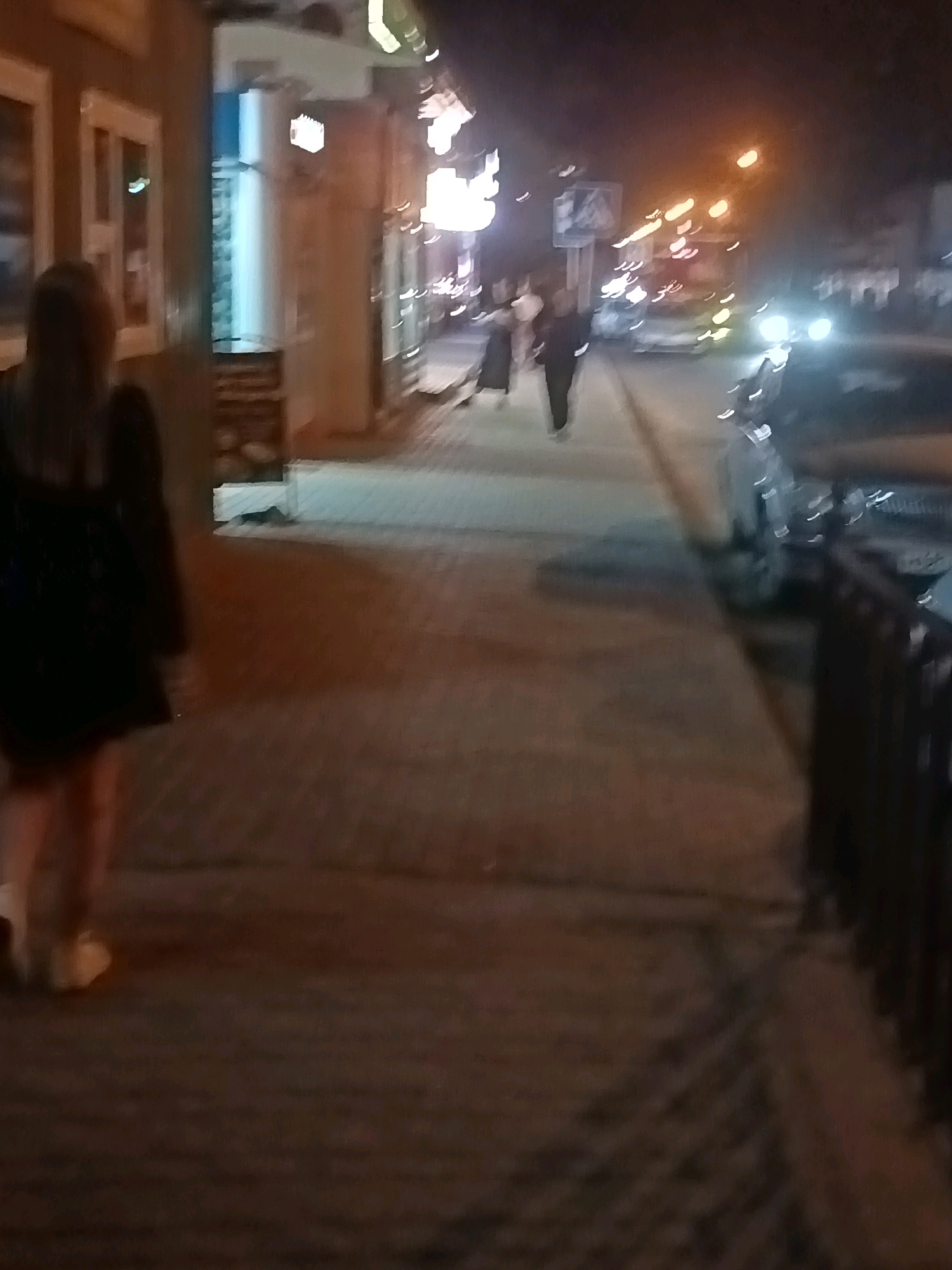 This is a nighttime, slightly blurry photograph of a street scene in Astrakhan, Russia. The image captures a sidewalk with a few people walking. On the left, a person with dark hair, wearing a dark dress and light-colored shoes, is walking away from the camera. Further down the sidewalk, two figures are walking towards the camera, one appearing to be wearing dark clothing. To the right of the sidewalk, cars are parked or driving by, with their headlights visible. Buildings with illuminated windows line the left side of the street. The overall atmosphere is dark, with artificial lights from street lamps and buildings creating streaks and reflections.