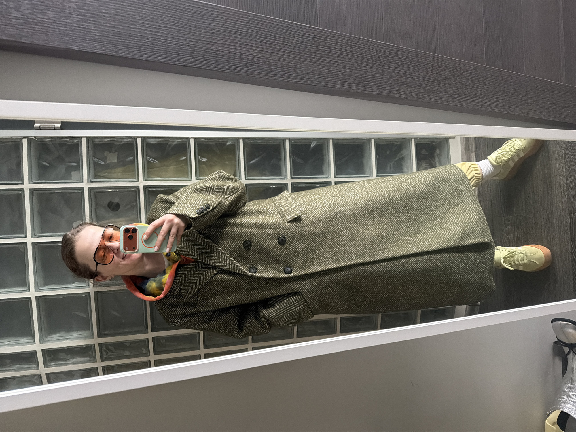 A person takes a mirrored selfie indoors. They are wearing orange-tinted glasses, a multicolored hoodie, and a patterned overcoat. The person is wearing light-colored sneakers with white socks. The background features a wall of glass blocks and wood paneling. The location cannot be determined from the image.