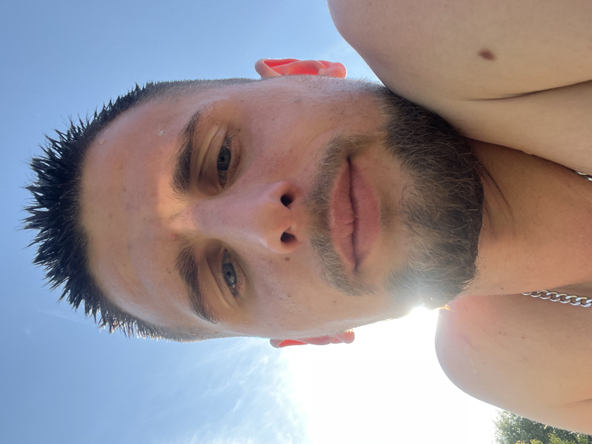 A male individual is depicted in a close-up, upward-facing selfie taken outdoors under a clear blue sky with scattered white clouds. The subject has short, dark, wet, spiked hair, a trimmed beard, and a mustache. He appears bare-chested, with a silver-colored chain visible on his left side. A dark, circular mole is present on his right shoulder. Strong sunlight originates from the lower-left, creating significant lens flare and backlighting the subject's left side, illuminating his wet hair and left ear. A small amount of green foliage is visible in the extreme lower-left corner. The individual is looking directly at the camera with a neutral expression. The location is Kapustino, Russia.