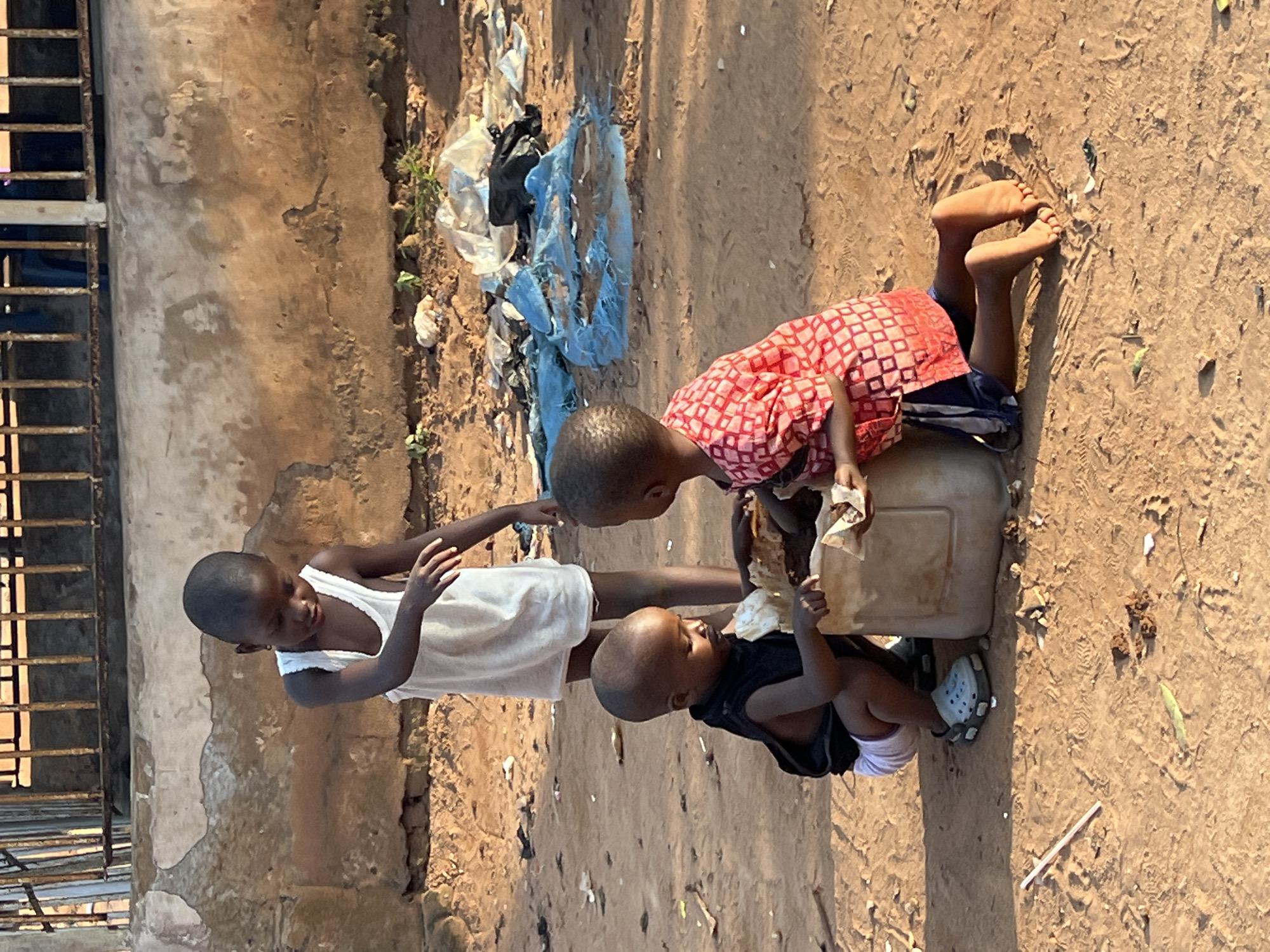 In Nkpor, Nigeria, three young children are gathered around a large, beige plastic container on the dusty ground. The eldest child, a boy wearing a white tank top and shorts, stands behind the container, reaching down towards it. To his left, a younger boy in a black sleeveless shirt and white shorts kneels, also reaching into the container. To the right, a third child, wearing a red and white patterned shirt and dark shorts, kneels and appears to be interacting with the contents of the container. The environment is characterized by a rough, uneven dirt surface littered with debris, including plastic bags and other discarded items. In the background, a weathered, textured wall with patches of peeling plaster is visible, topped by a rusty metal fence. The lighting suggests it is daytime, casting shadows on the ground.