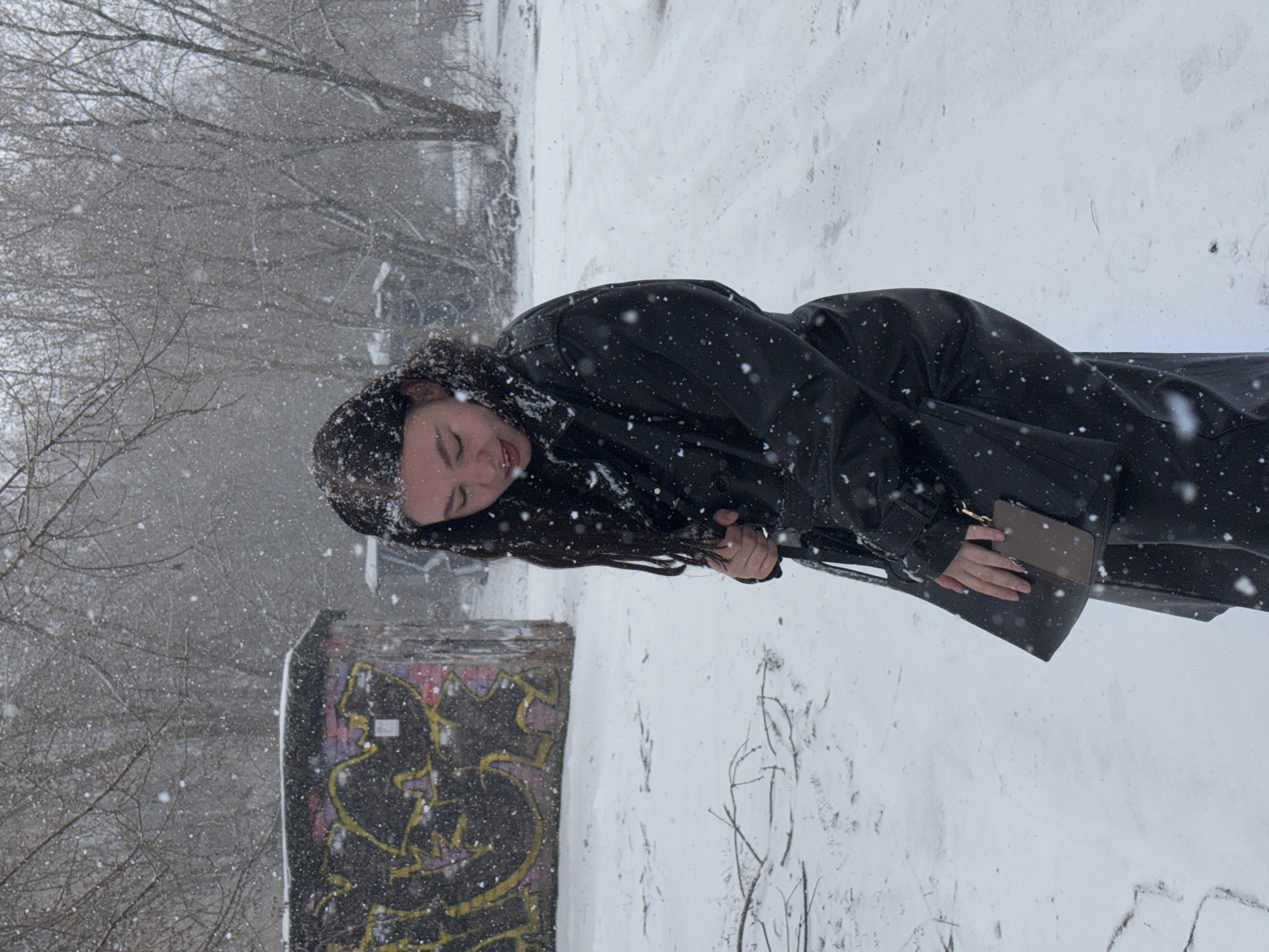 This image, taken in Perm, Russia, captures a young woman standing outdoors during a heavy snowfall. The scene is presented sideways, with a 90-degree clockwise rotation. When viewed upright, the woman is seen in profile from the chest up, her long, dark hair and black, possibly leather, long coat covered with numerous white snowflakes. Her head is tilted slightly down, eyes closed or cast downwards, and her mouth slightly agape, suggesting she is experiencing the falling snow on her face. Her left hand is visible, holding a small, light brown wallet or clutch.
The background depicts a stark winter environment. The ground is uniformly covered in thick white snow. To the left of the woman, a low, rectangular concrete structure, possibly a wall or barrier, is heavily covered in vibrant, unreadable graffiti in shades of black, yellow, and purple. Beyond this structure and in the upper left portion of the image, numerous bare tree branches are visible, blending into the hazy, snow-filled air. The air itself is dense with falling snowflakes, emphasizing the intensity of the winter weather. The overall impression is one of a cold, blustery, and serene winter day.