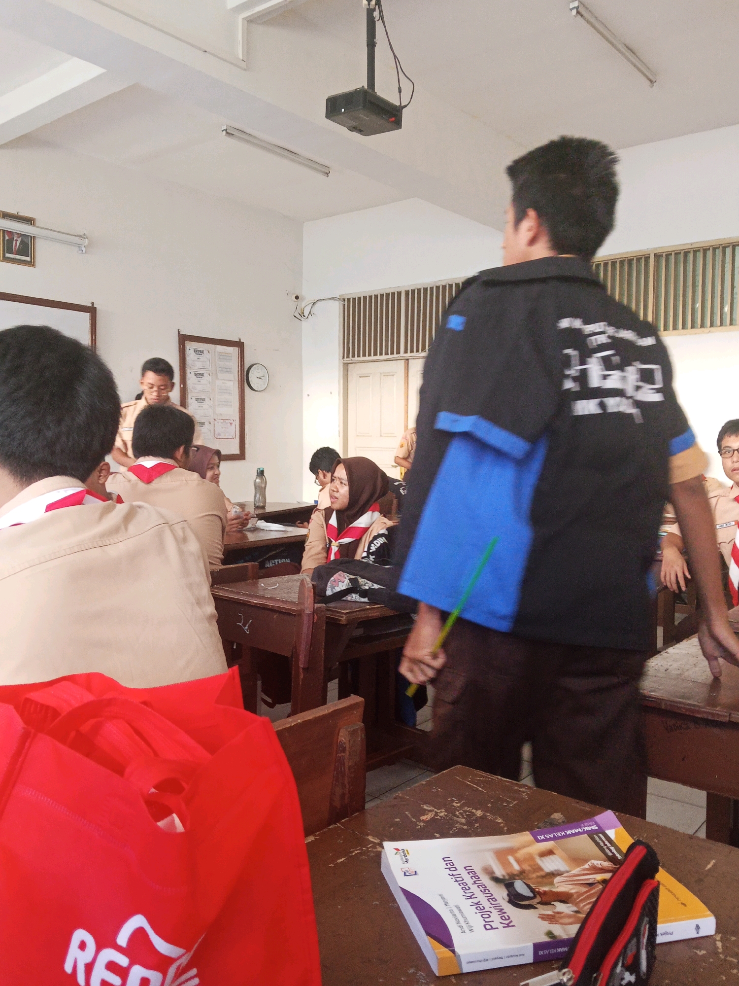 A group of Indonesian students, many in scout uniforms, are gathered in a classroom in Mangunjaya, Indonesia. A young man in a black shirt and brown pants stands facing away from the camera, holding a green stick. Other students are seated at desks, some wearing brown scout uniforms with red neckerchiefs and headscarves. A bulletin board with papers and a clock is mounted on the wall. Two red tote bags are visible in the foreground, and a book titled "Proyek Kreatif dan Kewirausahaan" is open on a desk. The room appears to be brightly lit with overhead fluorescent lights.