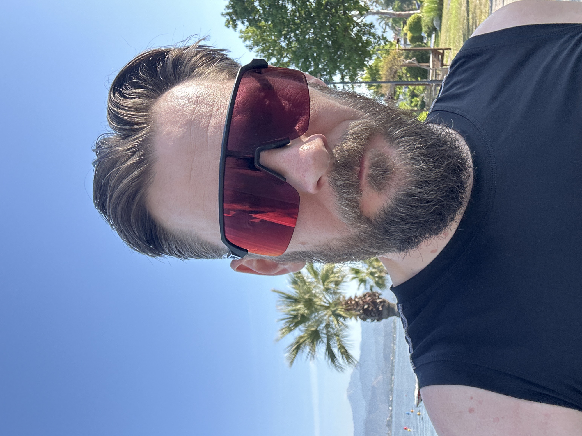 A rotated close-up shot captures a man outdoors on a bright, sunny day. The man has short, dark brown hair, which appears slightly windswept, and a thick, dark beard with hints of grey, covering his chin and cheeks. He is wearing large, frameless, red-tinted wrap-around sunglasses, obscuring his eyes and a significant part of his face. His attire consists of a simple black short-sleeved athletic top or t-shirt.
The background reveals a vibrant clear blue sky, indicating excellent weather conditions. Scattered green foliage is visible, including a prominent palm tree and other distant trees. In the mid-ground, a body of water, likely the sea, is visible with distant mountains or landforms on the horizon. Several small, colorful objects, possibly buoys or water toys, are floating in the water near the foreground, suggesting a recreational area.
The overall scene suggests a leisure activity or vacation in a warm, coastal environment during the daytime. Given the context, the location is Fethiye, Turkey. There is no visible text in the image.
