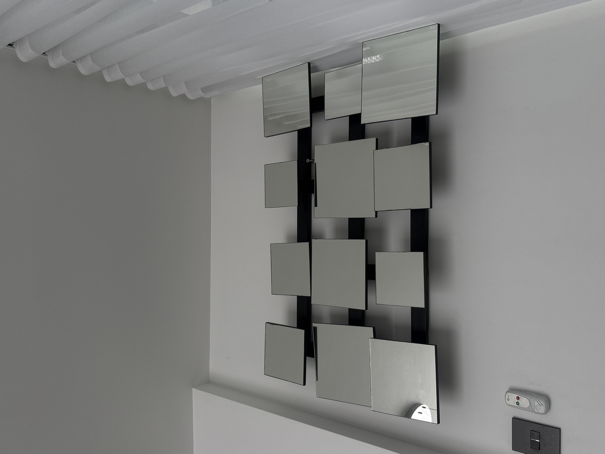 A modern wall decoration is seen in a room with gray walls, potentially located in a home or office space in San Bernardino Tlaxcalancingo, Mexico. The wall decoration consists of multiple square mirrors arranged in a grid-like pattern, mounted on a black frame. The mirrors reflect the surrounding environment, creating an interesting visual effect. Below the wall decoration is a white baseboard, and on the wall, to the right of the mirrors, are a remote control device and a light switch. The ceiling is adorned with white, sheer curtains. The room appears to be well-lit and clean, with a minimalist aesthetic.
