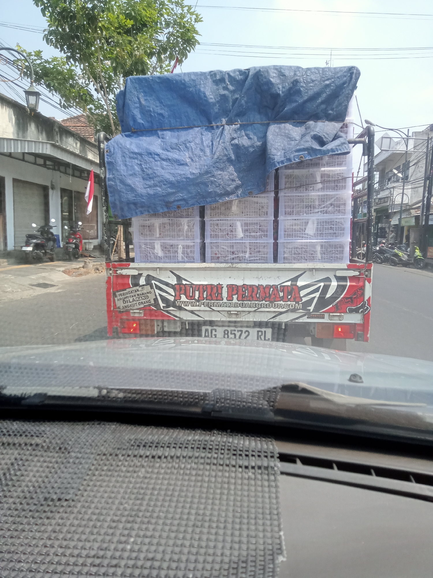 This is a street-level, eye-level, medium shot of a red and white pickup truck with a blue tarp covering its cargo, driving on a street in Mangunsari, Indonesia. The truck is loaded with white plastic crates, possibly containing goods. The truck has markings on its rear that include "PUTRI PERMATA" and a website address. Below that, a license plate reads "AG 8572 RL". To the left of the truck, there are buildings with closed storefronts and parked motorcycles. A red and white Indonesian flag is visible on one of the buildings. The sky is clear and blue, with visible power lines crisscrossing above. The perspective is from inside another vehicle, looking out through the windshield, which shows part of the dashboard in the foreground. The overall scene depicts a typical urban street environment in Indonesia.