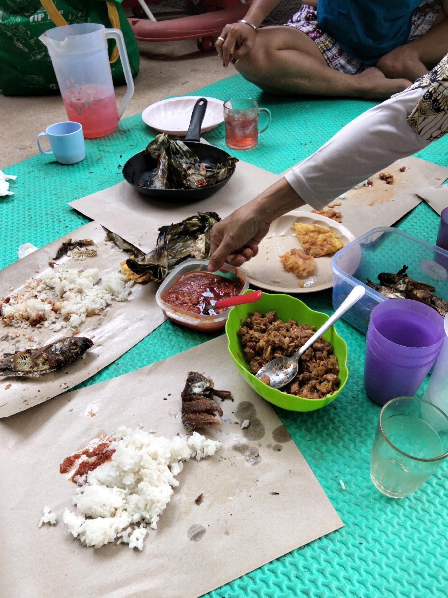 In Rawa Atug, Indonesia, a casual outdoor meal is in progress. The scene unfolds on a green mat, with several individuals seated around a spread of food served on brown paper. The fare includes white rice, fried fish, and a dish of what appears to be crumbled tempeh or fried minced meat. A small bowl of red chili paste, likely sambal, is also present.
A hand, adorned with a ring, is captured reaching towards a container of sambal, suggesting an active participation in serving or eating. Nearby, a clear pitcher holds a pinkish beverage, accompanied by a small blue mug. A clear glass contains a light-colored liquid. Two purple plastic cups, one empty and one partially filled, sit alongside a transparent plastic container holding more fried fish. The setting appears informal, with the food laid out on the ground, indicative of a communal dining experience. The backdrop suggests an outdoor location, possibly a yard or park, with elements of what looks like outdoor furniture or equipment visible in the upper portion of the image.