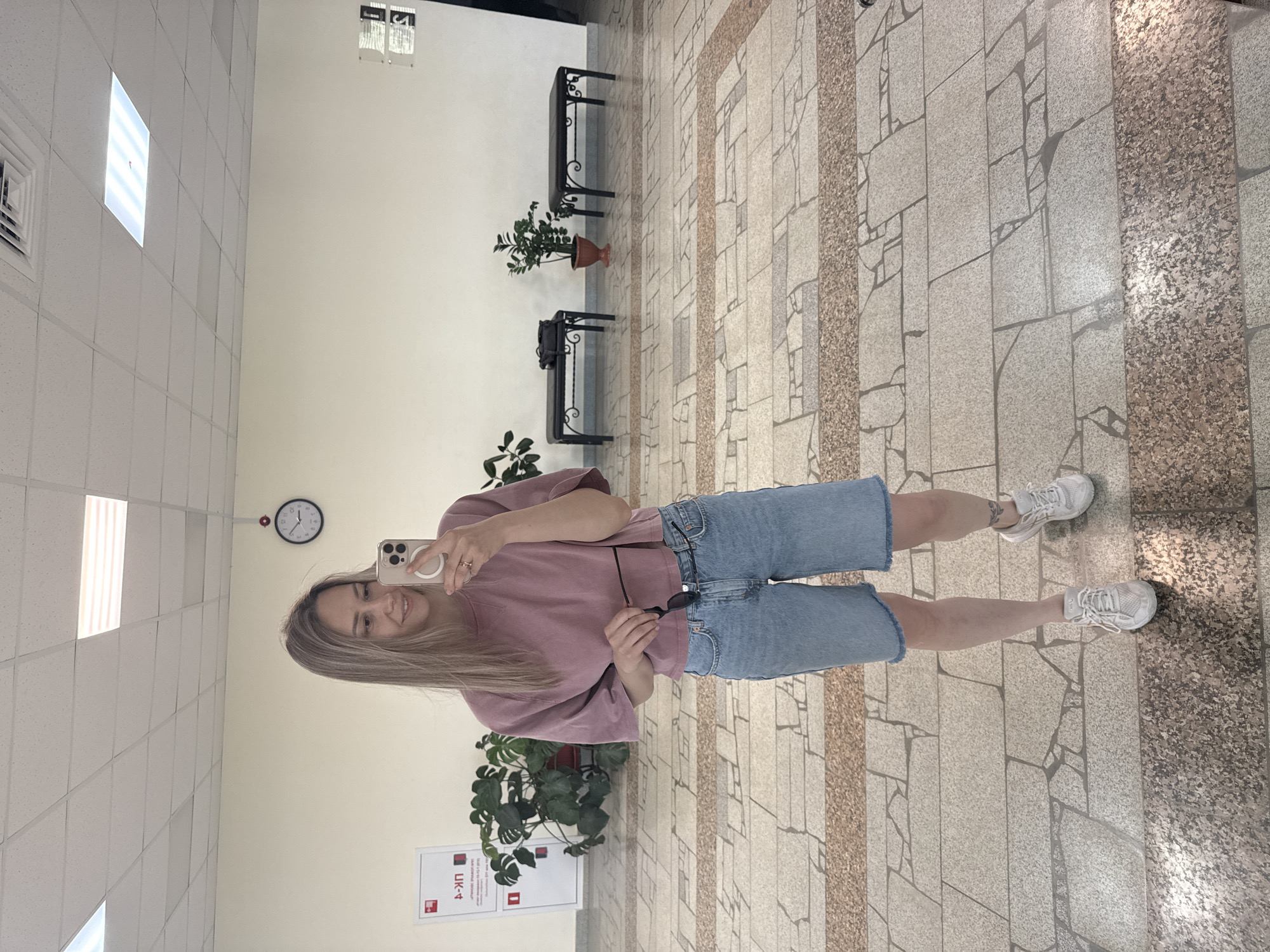 A woman stands in a corridor, taking a selfie. She has blonde hair, wears a pink t-shirt and blue denim shorts, and white sneakers. She holds a cell phone in her right hand. Sunglasses dangle from her left hand. In the background are indoor plants in pots, a clock on the wall, and benches along the wall. The floor is tiled. A sign with "ЦК-4" is visible in the lower-left corner. Based on this sign and location information, the picture may be taken in Noviy Urengoy, Russia.