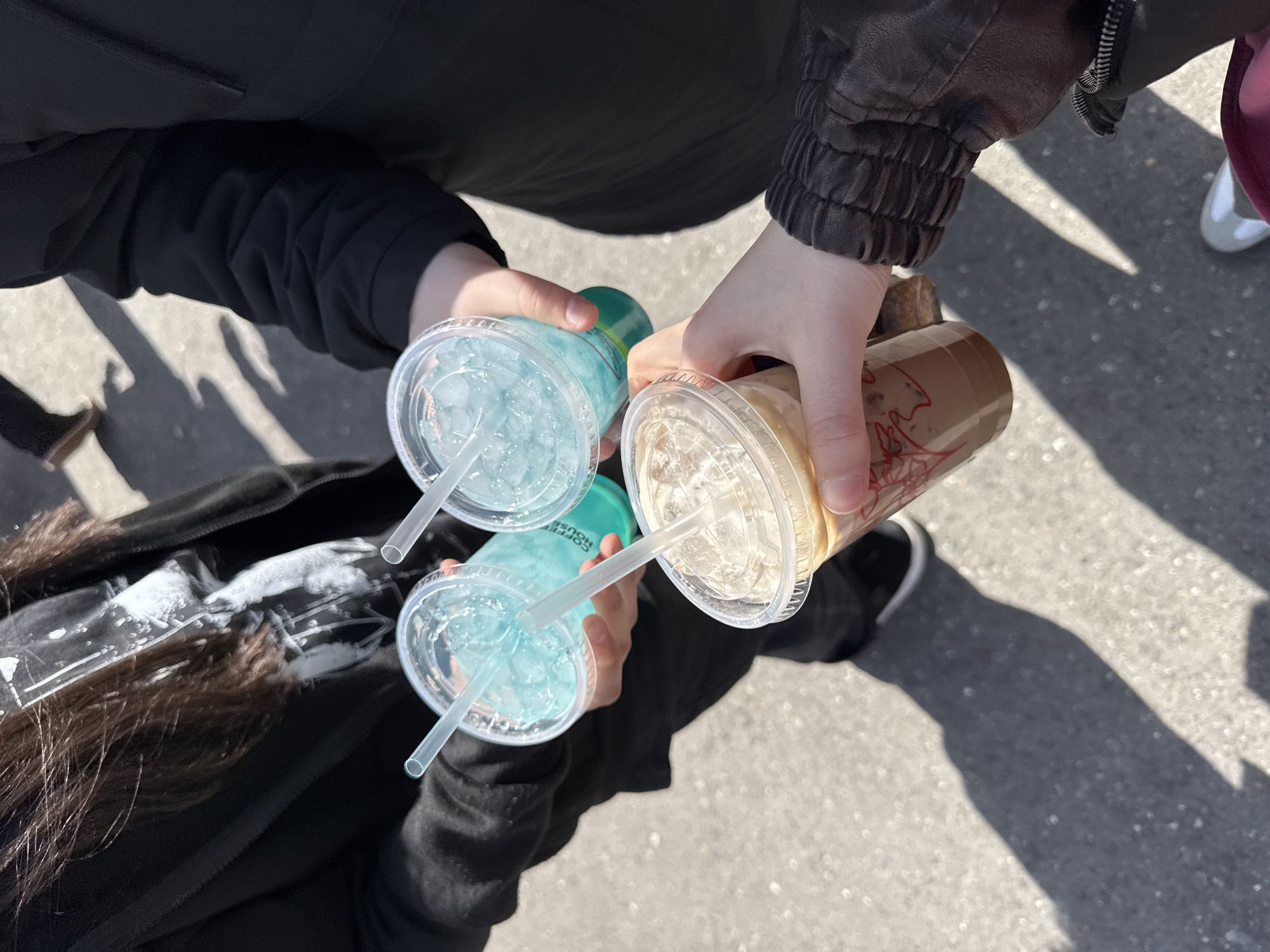 Three people are holding beverages in plastic cups on a paved surface. Two cups contain a blue liquid with ice, and one contains a brown liquid. Two of the people are wearing black jackets, and one has dark hair with blonde highlights. A white shoe can be partially seen in the upper right corner. The location is unknown, and there is no evidence to suggest it is Yerevan, Armenia.