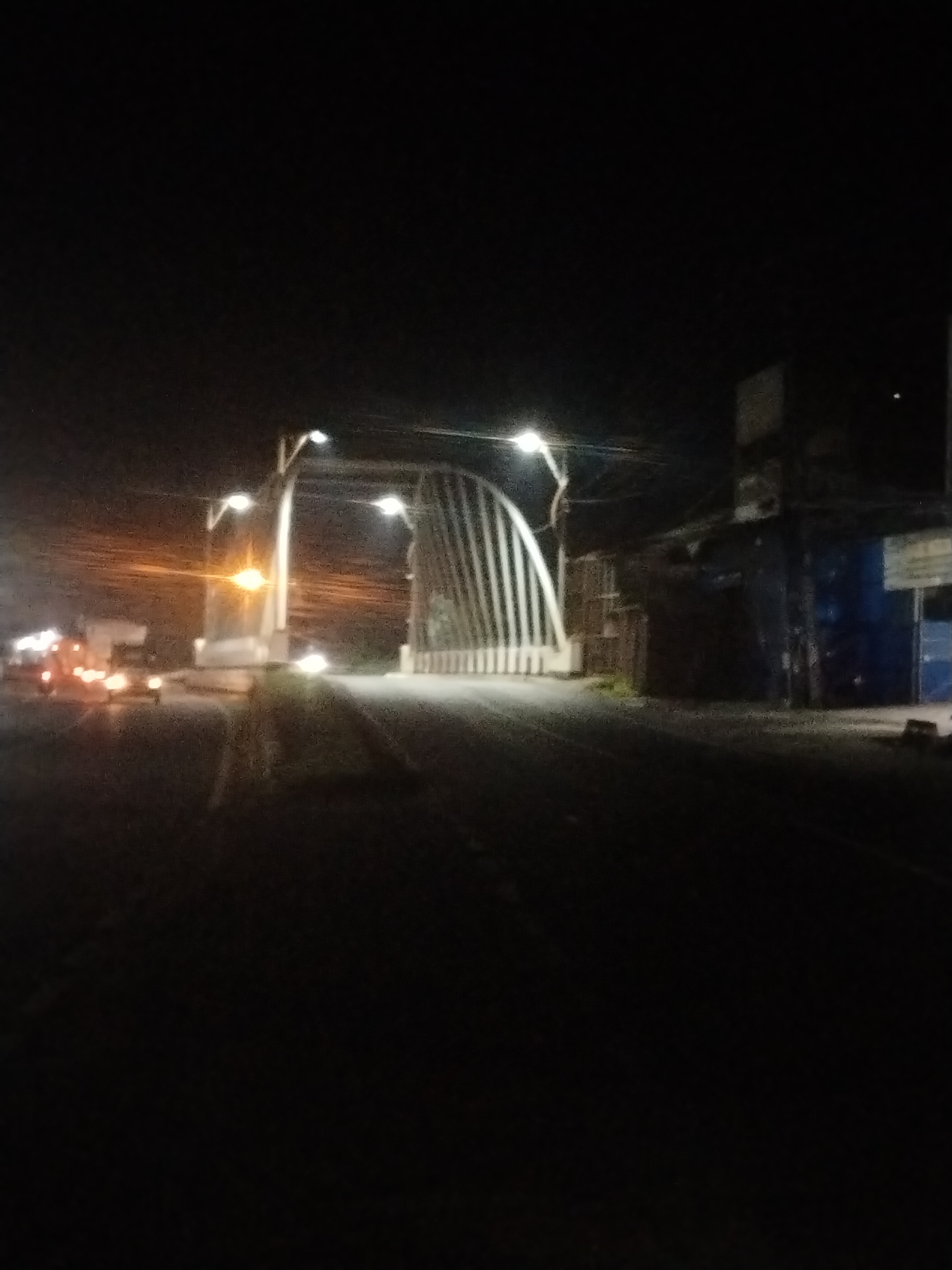 At night in Lhokseumawe, Indonesia, a street stretches toward an arched structure illuminated by overhead lights. Vehicles with lit headlights and taillights are present in the distance, on the left side of the road. To the right of the arch, a building with dark colors and signage is visible. The foreground of the street is dark and indistinct.
