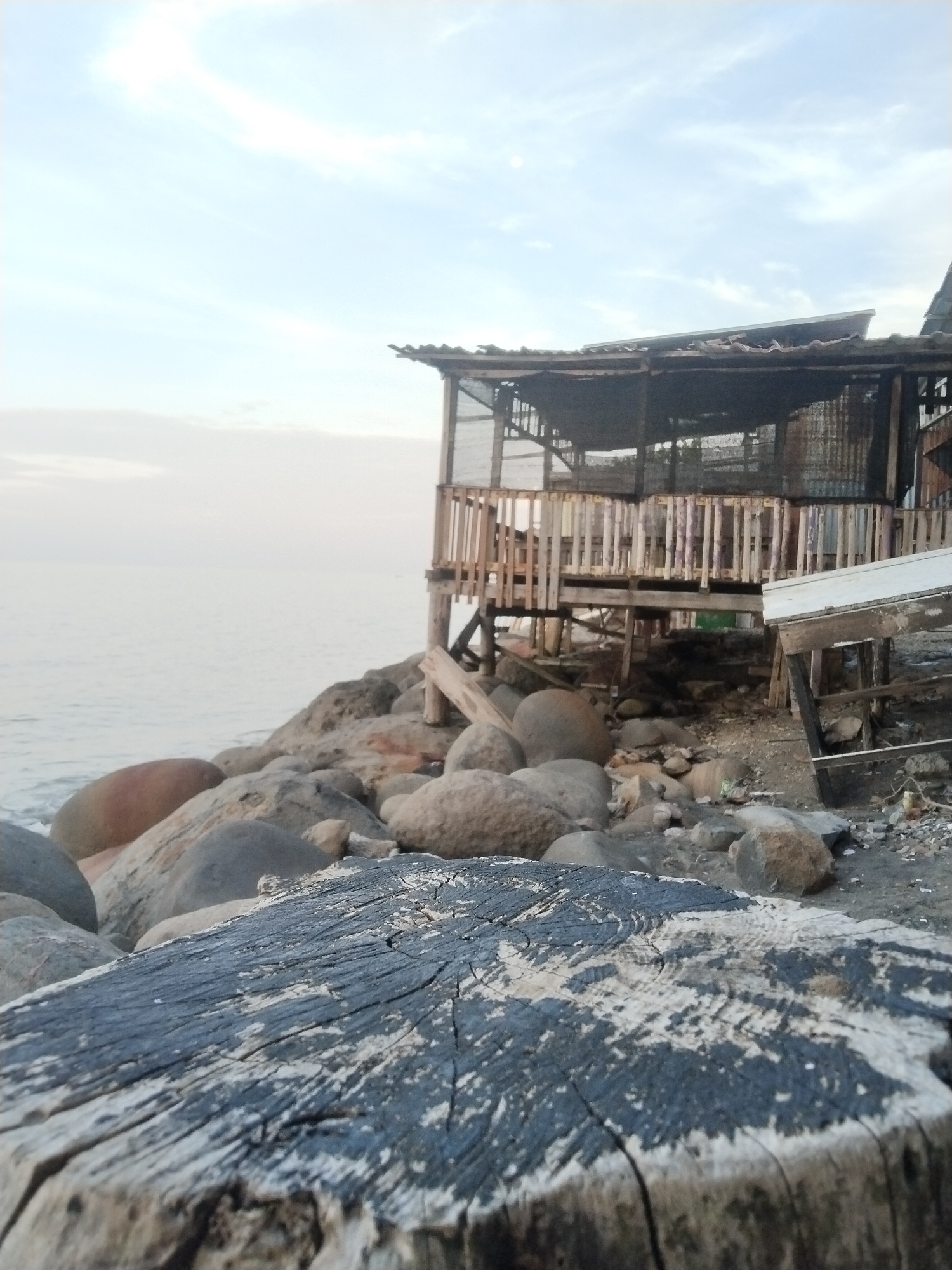 This image depicts a tranquil coastal scene in Lhokseumawe, Indonesia, likely captured during the soft light of either early morning or late afternoon. The sky above is a pale blue with faint, wispy clouds, suggesting calm weather.
In the foreground, a prominent, weathered wooden stump dominates the lower part of the frame. Its surface reveals intricate wood grain patterns and areas where dark and white textures, possibly old paint or natural discoloration, contrast sharply.
Behind the stump, a rocky shoreline is visible, strewn with numerous large, smooth, rounded stones varying in size and earthy hues. Perched on these rocks, a rustic, elevated wooden structure stands on stilts. This structure, possibly a hut or small building, features wooden railings that appear worn and aged, and a dark roof which seems to be made of corrugated material or tarp. Parts of the structure are enclosed with a dark mesh or screen. Its overall appearance suggests a simple, functional construction that has endured the elements.
To the left of the structure, the calm, light-colored ocean extends serenely towards the horizon, merging with the soft sky. No people or specific activities are observed, contributing to the quiet, serene atmosphere of the coastal setting. There is no visible text within the image.