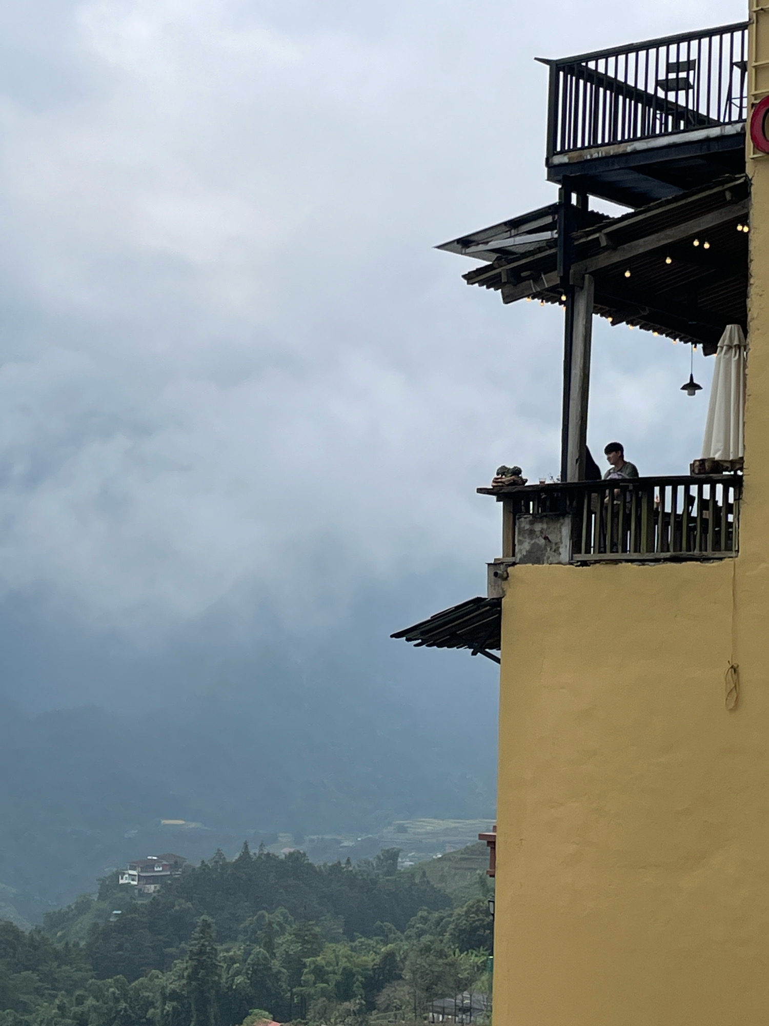 A medium shot captures a person standing on a balcony overlooking a misty, mountainous landscape in Sa Pa Ward, Vietnam. The balcony, constructed with dark wooden railings and a tiled roof, juts out from a yellow building on the right side of the frame. Lights are strung along the underside of the roof, and a white umbrella is partially visible. A person, wearing a dark shirt and pants, stands near the railing, facing away from the camera and towards the view. Below the balcony, the lush, green hills of Sa Pa are shrouded in a thick, grey mist, with glimpses of houses and terraced fields visible through the haze. The sky is overcast, contributing to the diffused light and atmospheric mood of the scene.
