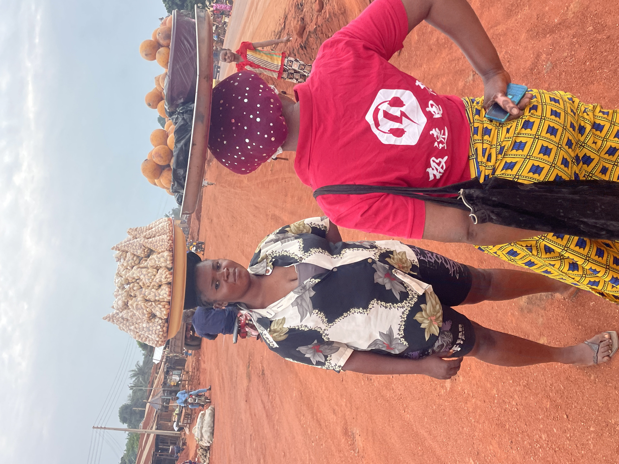 The image depicts a bustling street scene in Otukpa, Nigeria. Two women are prominently featured, each carrying goods on their heads. The woman in the foreground wears a black and white floral shirt and shorts, balancing a tray piled high with what appear to be nuts or seeds. The second woman, behind her, is dressed in a red t-shirt with a white logo, a patterned yellow and blue skirt, and a polka-dot headscarf. She also has a cell phone in her hand and carries a tray on her head, filled with round, orange-colored fruits. The ground is a reddish-brown color, indicative of the local soil. In the background, other figures and vehicles can be seen, suggesting a busy market or commercial area. The sky is overcast.