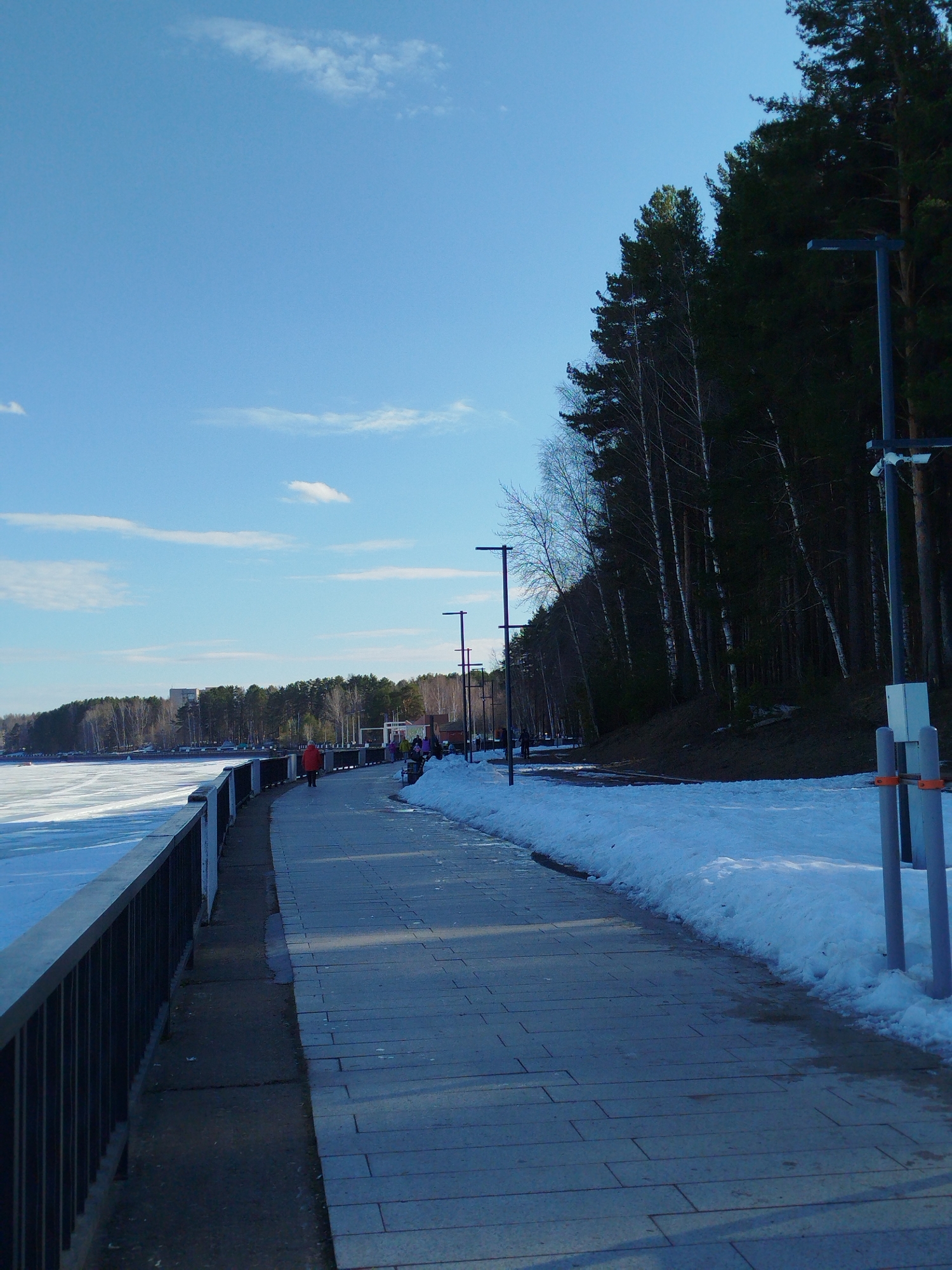 The image shows a paved pathway in Chaikovsky, Russia, alongside a partially frozen body of water, presumably the Kama River. A black metal railing separates the path from the icy water. Patches of snow linger on the ground beside the pathway, which is bordered by a line of trees and lamp posts. Several people are walking along the path. The sky is blue with scattered clouds.