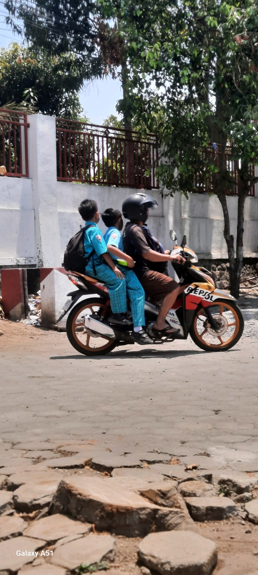 An adult rides a motorcycle carrying two children on a street in Tangkil, Indonesia. The adult, wearing a dark shirt, brown shorts, and a black helmet, is driving the motorcycle. The two children, dressed in blue school uniforms with checkered pants, sit behind the driver. The child in the middle has a black backpack. The motorcycle has orange and yellow decals with the word "REPSOL" visible. The street appears to be paved and cracked, with some debris on the side. Trees and a white wall with a red fence are visible in the background, suggesting a residential or school area. The lighting indicates it is daytime, with sunlight casting shadows.