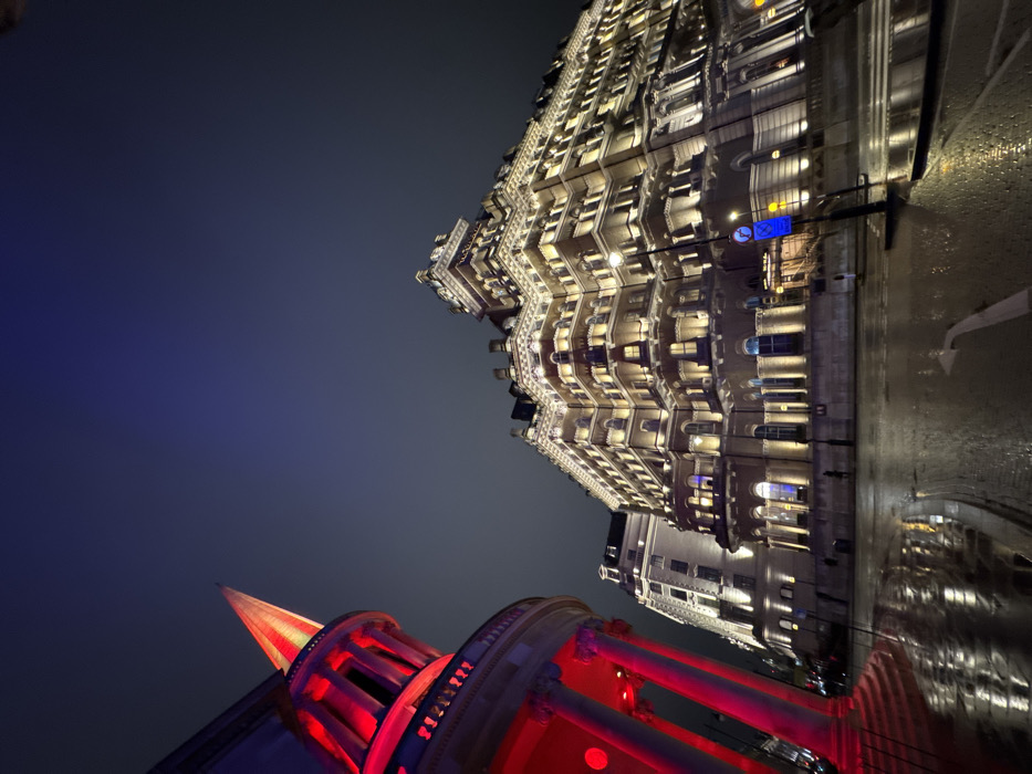 Low-angle shot of two buildings at night. One is lit with red lights, featuring a pointed spire. The other is a large, ornate building illuminated with white lights, multiple stories high. The street reflects the building's lights, and a blue sign is visible. The sky is a gradient of dark blue.