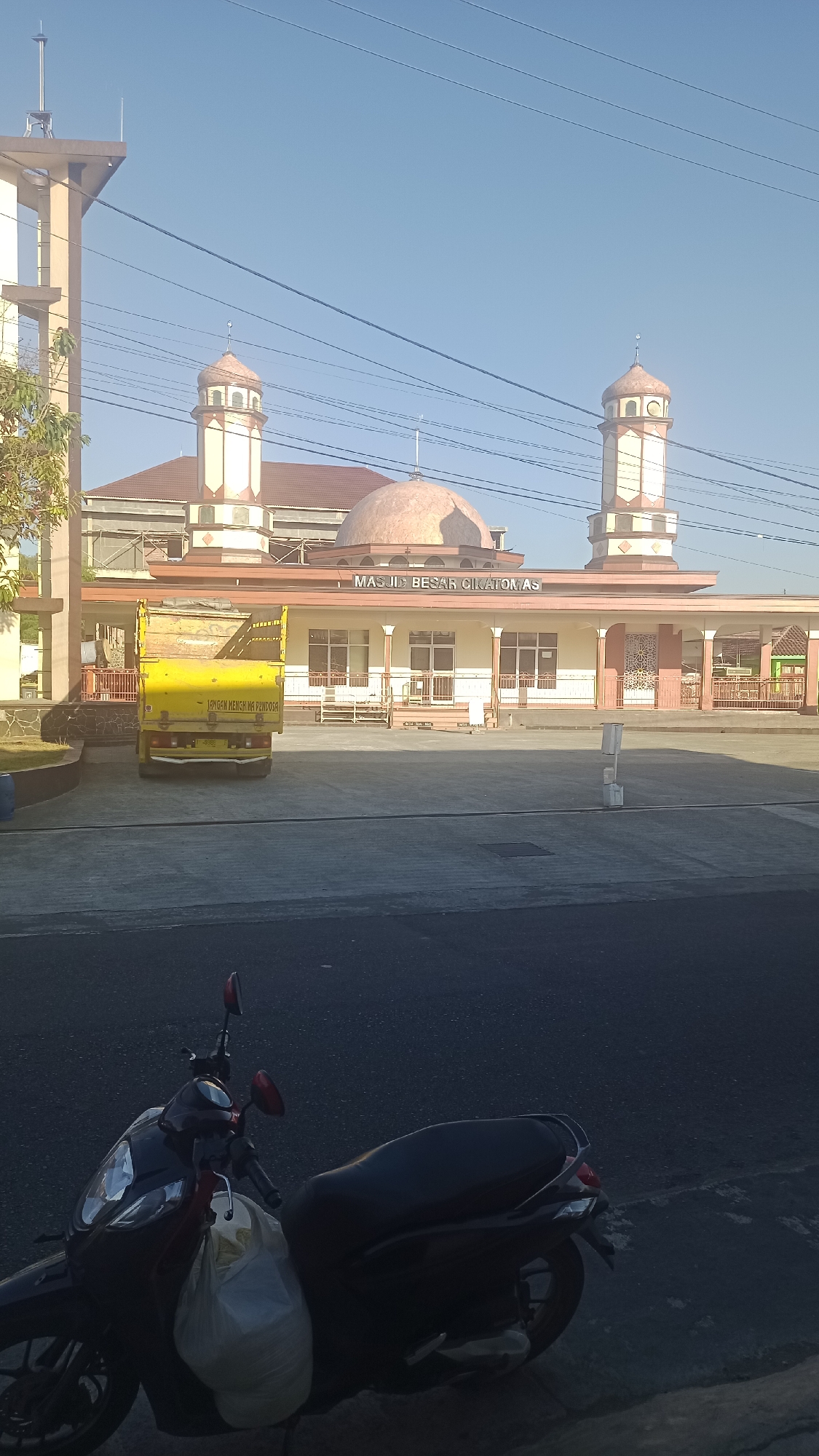 The image shows a mosque with two minarets and a large dome. The mosque is named "MASJID BESAR CIKSATOMAS". There is a yellow truck parked in front of the mosque, and a scooter is parked in the foreground. The sky is clear and blue, and the scene appears to be set in a city. The location is Pakemitan, Indonesia.