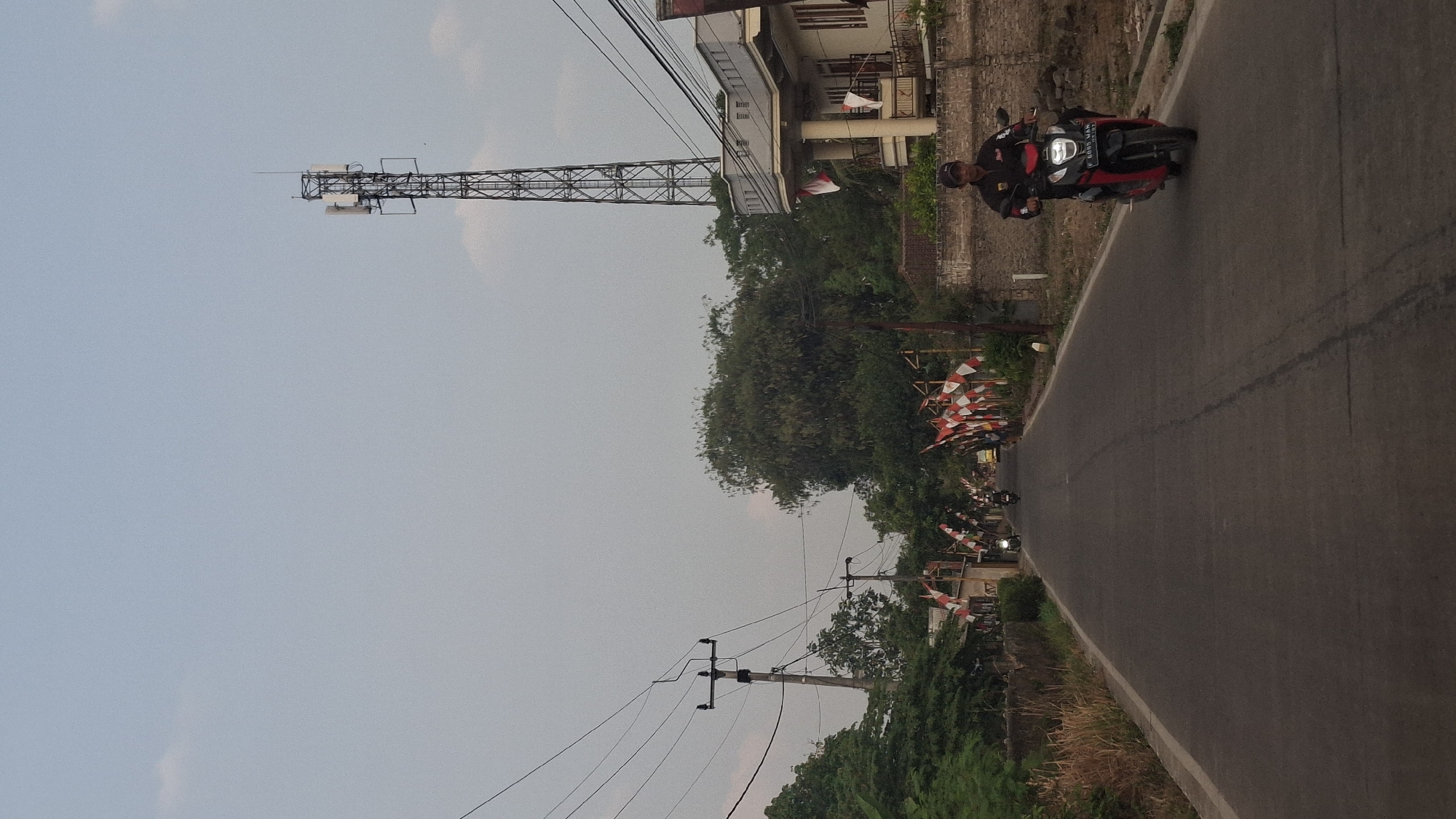In Cibinong Hilir, Indonesia, a rural road stretches forward under an overcast sky. On the right side of the road, a man on a red and black motorcycle, wearing a black jacket and a cap, rides towards the viewer. Behind him, a beige building with a covered porch and a balcony stands, adorned with Indonesian flags. Further down the road, more flags are visible, lining the street. A tall telecommunications tower dominates the skyline on the right. To the left of the road, a power pole with electrical wires is present, along with lush green trees and vegetation. Several other motorcycles and a yellow vehicle are seen further up the road, receding into the distance.