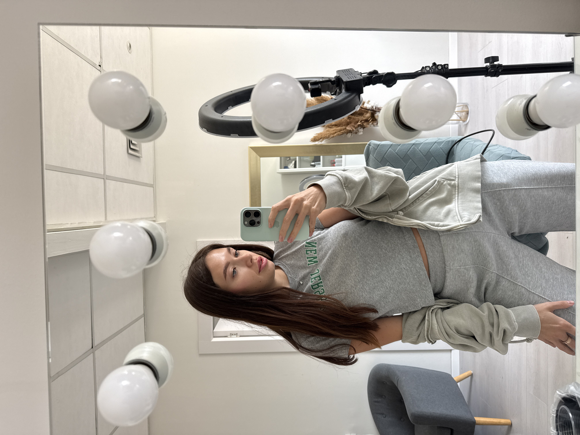 A person stands in front of a mirror in a brightly lit interior. The mirror is surrounded by evenly spaced white light bulbs. The person is a young woman with long, dark brown hair, wearing a gray sweatsuit. She is holding a light green cell phone, apparently taking a selfie. A ring light on a stand is positioned behind her, visible in the mirror's reflection. In the background, there is a light blue upholstered chair, a gray chair, and another mirror reflecting a shelf with items on it. The location cannot be determined based on the image, so Ramat Gan, Israel cannot be confirmed.