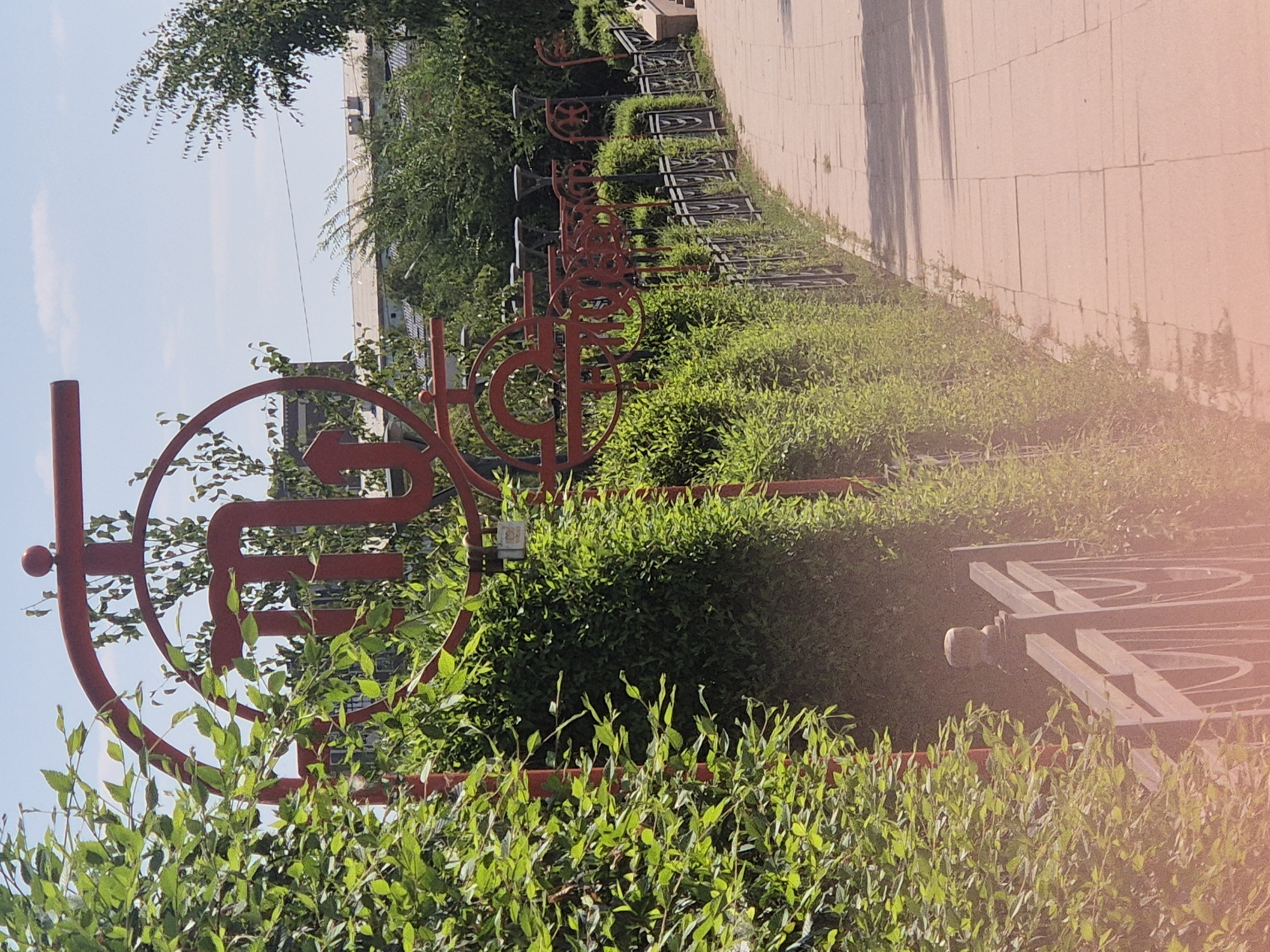 The image depicts a pathway in Kokshetau, Kazakhstan, lined with manicured hedges and a decorative metal fence. Prominently featured on the left is a large, red, abstract metal sculpture resembling a stylized scorpion with an arrow pointing upwards. Further along the pathway, a series of similar red metal decorative elements are visible, interspersed with lampposts. The pathway itself is paved with square concrete tiles. In the background, buildings and trees are discernible under a clear blue sky. The overall scene suggests a well-maintained public space, possibly a park or a promenade, in a city setting.