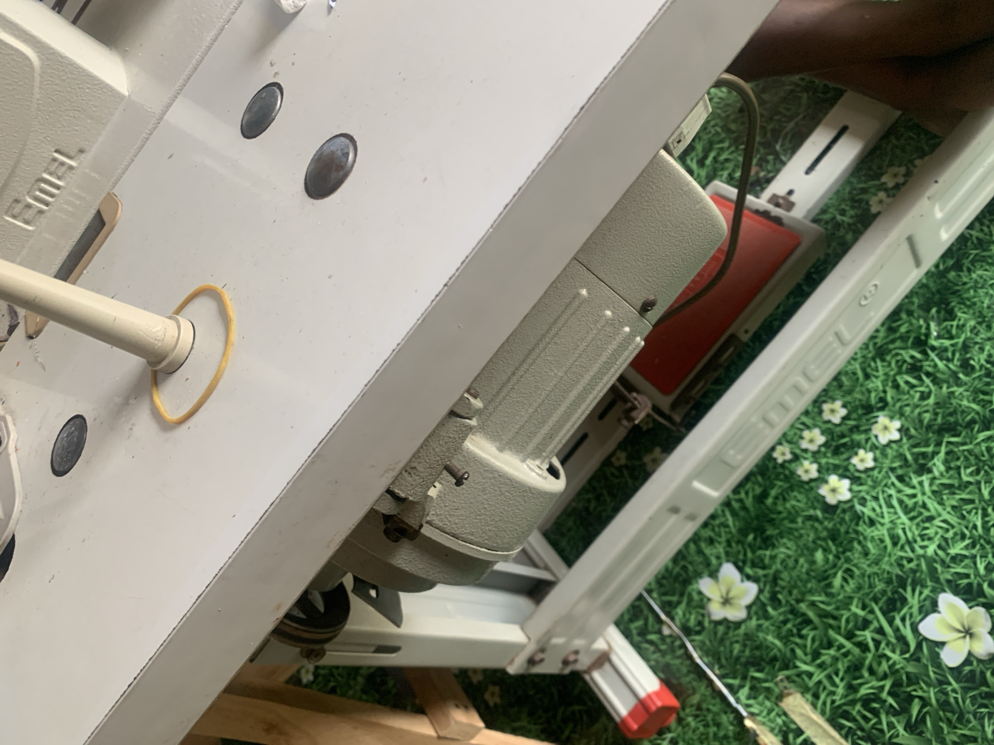 This image depicts a close-up, angled view of a white industrial sewing machine, possibly located in Jauro Yinu, Nigeria. The sewing machine table is the dominant surface, appearing clean with scattered debris. A beige motor is attached to the underside of the table, connected by a belt to a pulley system. A yellow rubber band is visible on the table surface. The machine has embossed branding that reads "Emel". The sewing machine is situated over a green, artificial grass-like mat that is patterned with small white flowers. The background is out of focus, but appears to be a wooden structure. No people are visible in the frame.